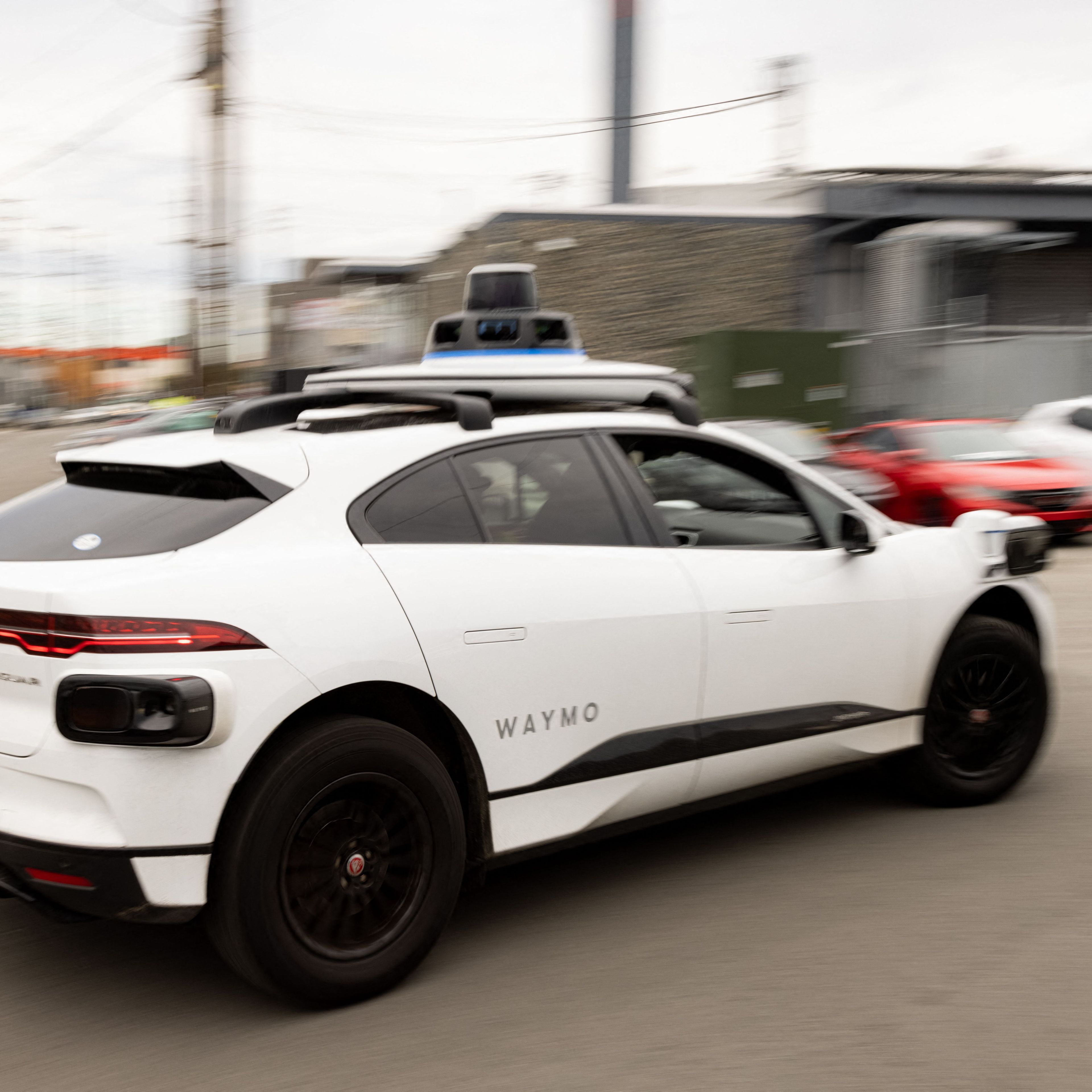 A white Waymo-branded self-driving car with sensors on the roof is driving on a street with several parked cars and industrial buildings in the background.