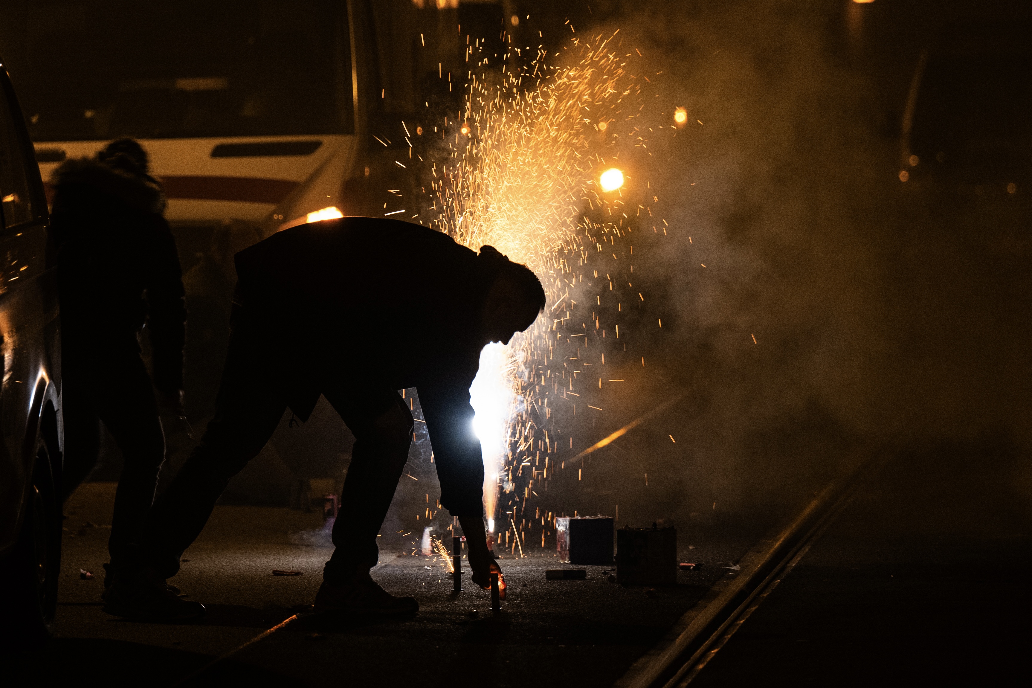 A person bends down to light fireworks in the street at night, with sparks flying and smoke filling the air, illuminating the otherwise dark surroundings.