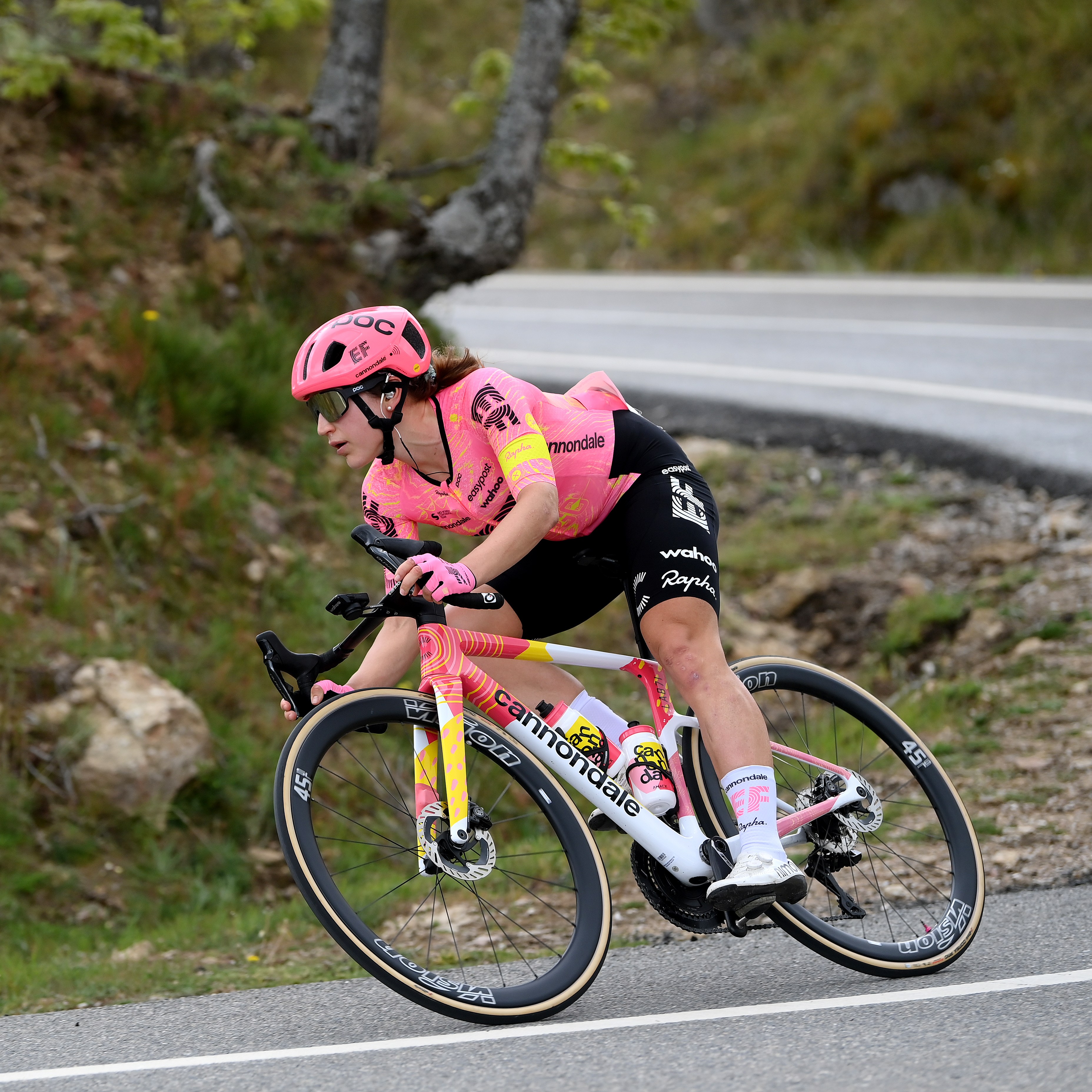 A cyclist wearing a pink jersey and helmet is riding a white Cannondale bike around a curve on a road, with greenery and trees in the background.