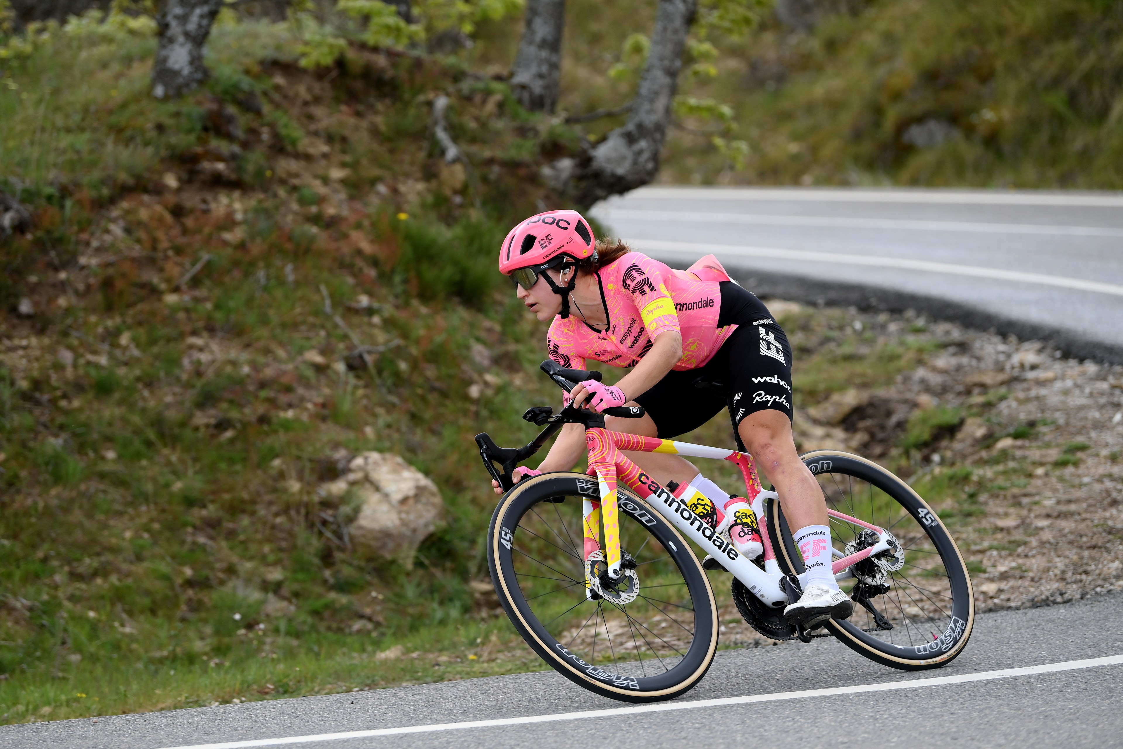 A cyclist wearing a pink jersey and helmet is riding a white Cannondale bike around a curve on a road, with greenery and trees in the background.