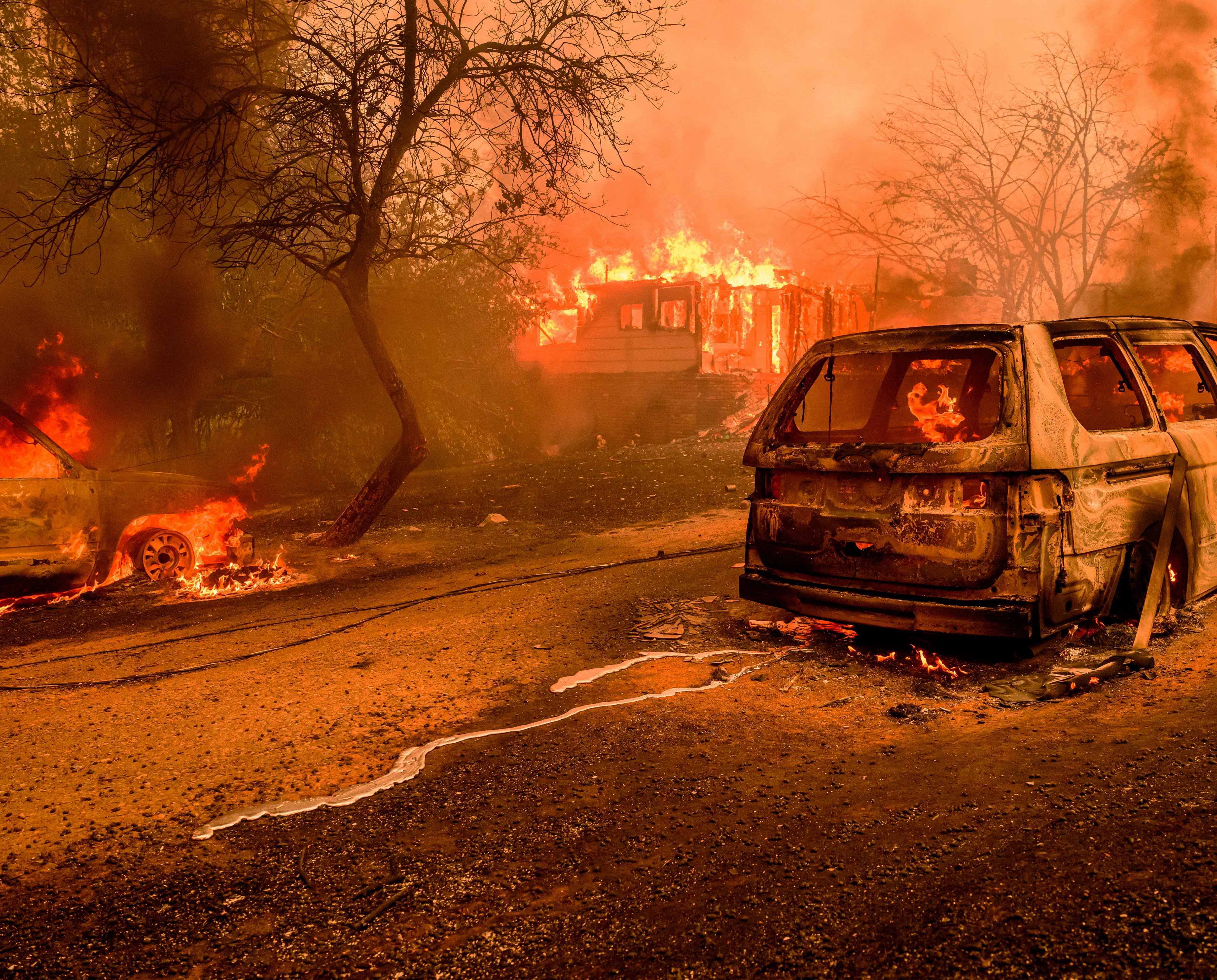 The image shows a devastated scene with two burning cars on a road, a house engulfed in flames, and charred trees under an orange sky filled with thick smoke.