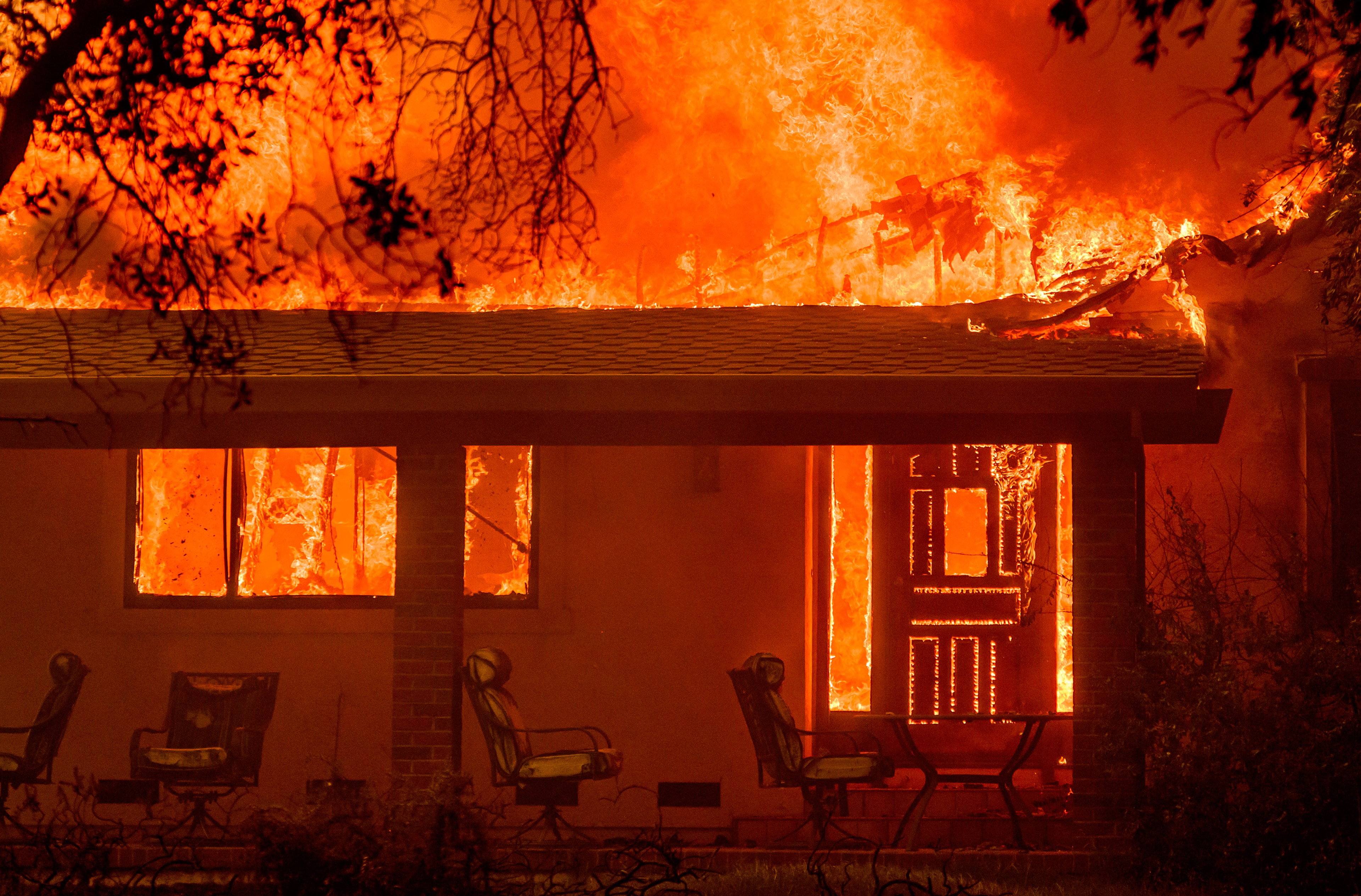 A house is engulfed in intense flames, with the roof and windows ablaze. Outdoor furniture is visible on a porch which has not yet caught fire. Tree branches frame the scene.