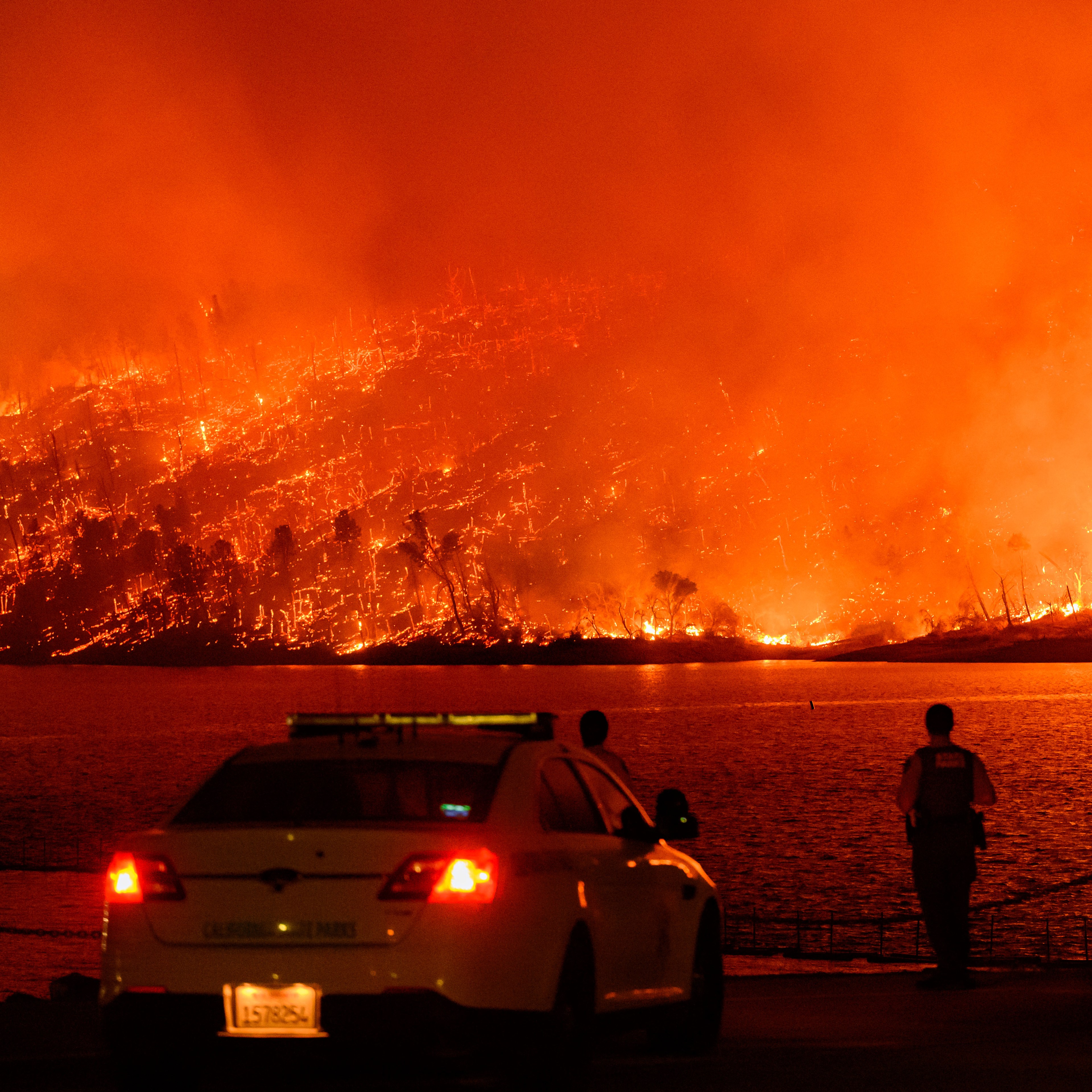 A massive wildfire engulfs a hillside, casting an orange glow across the sky and reflecting off a body of water. Emergency personnel and a vehicle are in the foreground.