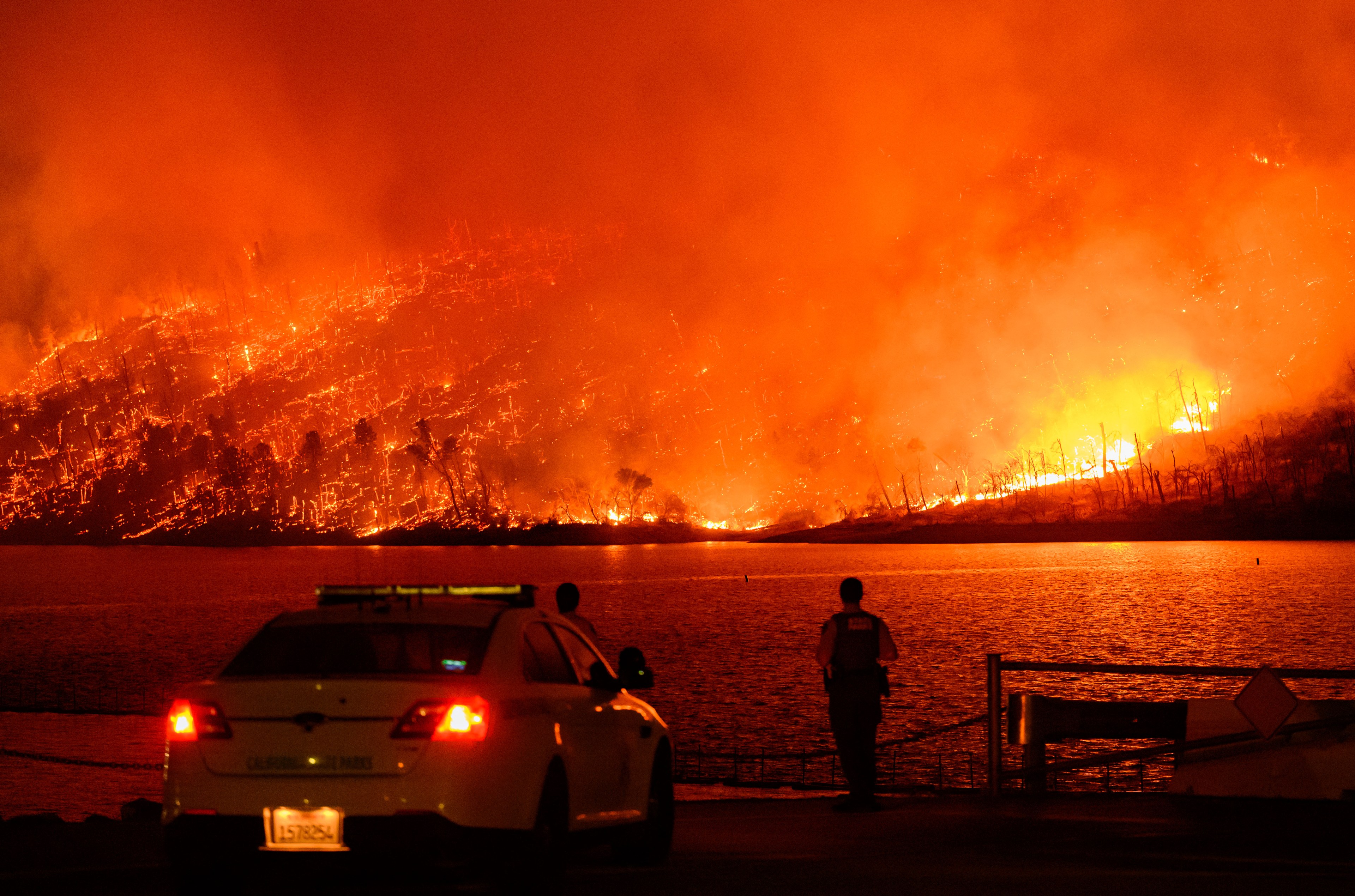 A massive wildfire engulfs a hillside, casting an orange glow across the sky and reflecting off a body of water. Emergency personnel and a vehicle are in the foreground.
