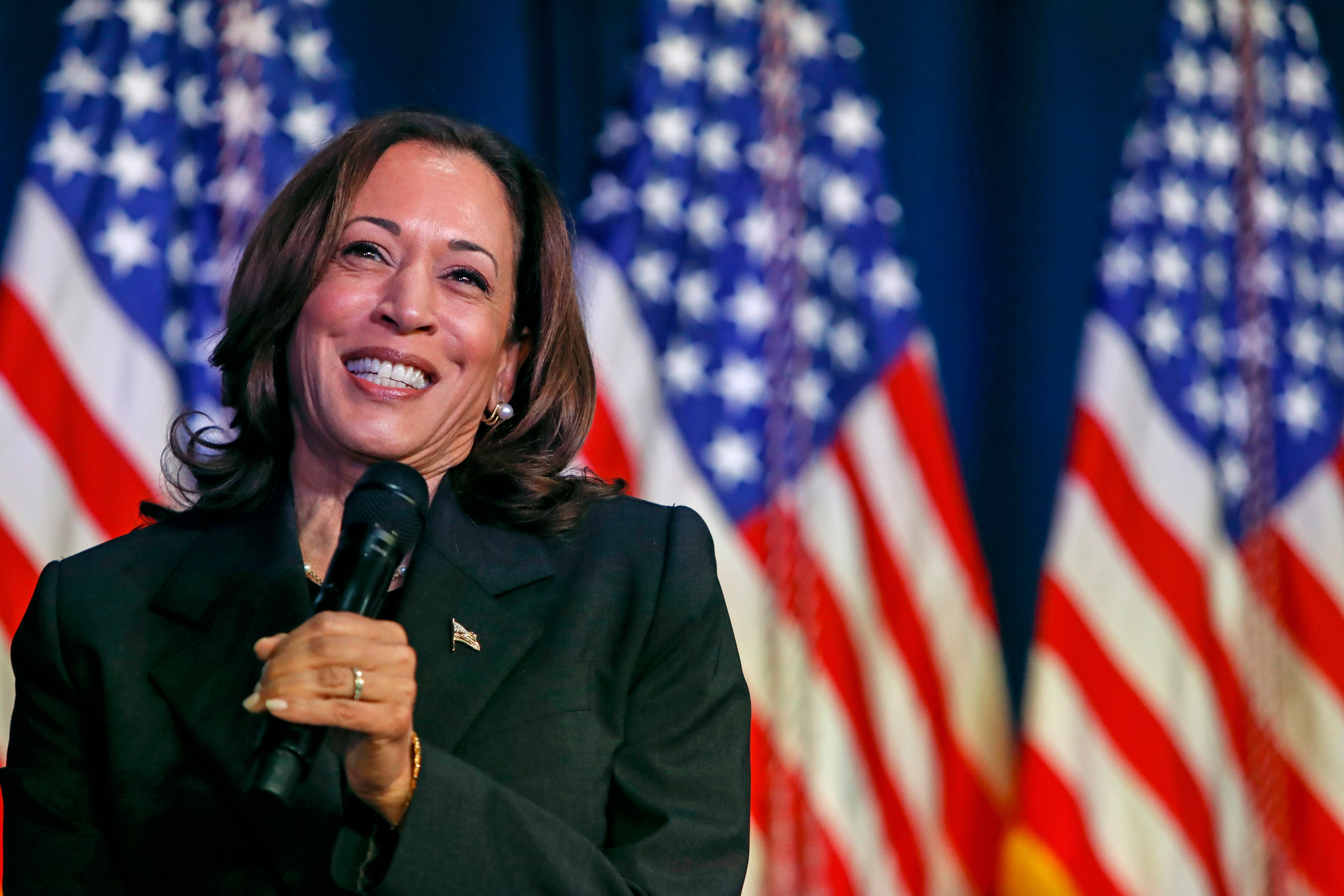 A smiling woman holds a microphone and speaks at an event, with several out-of-focus American flags in the background. She wears a dark suit and has shoulder-length hair.