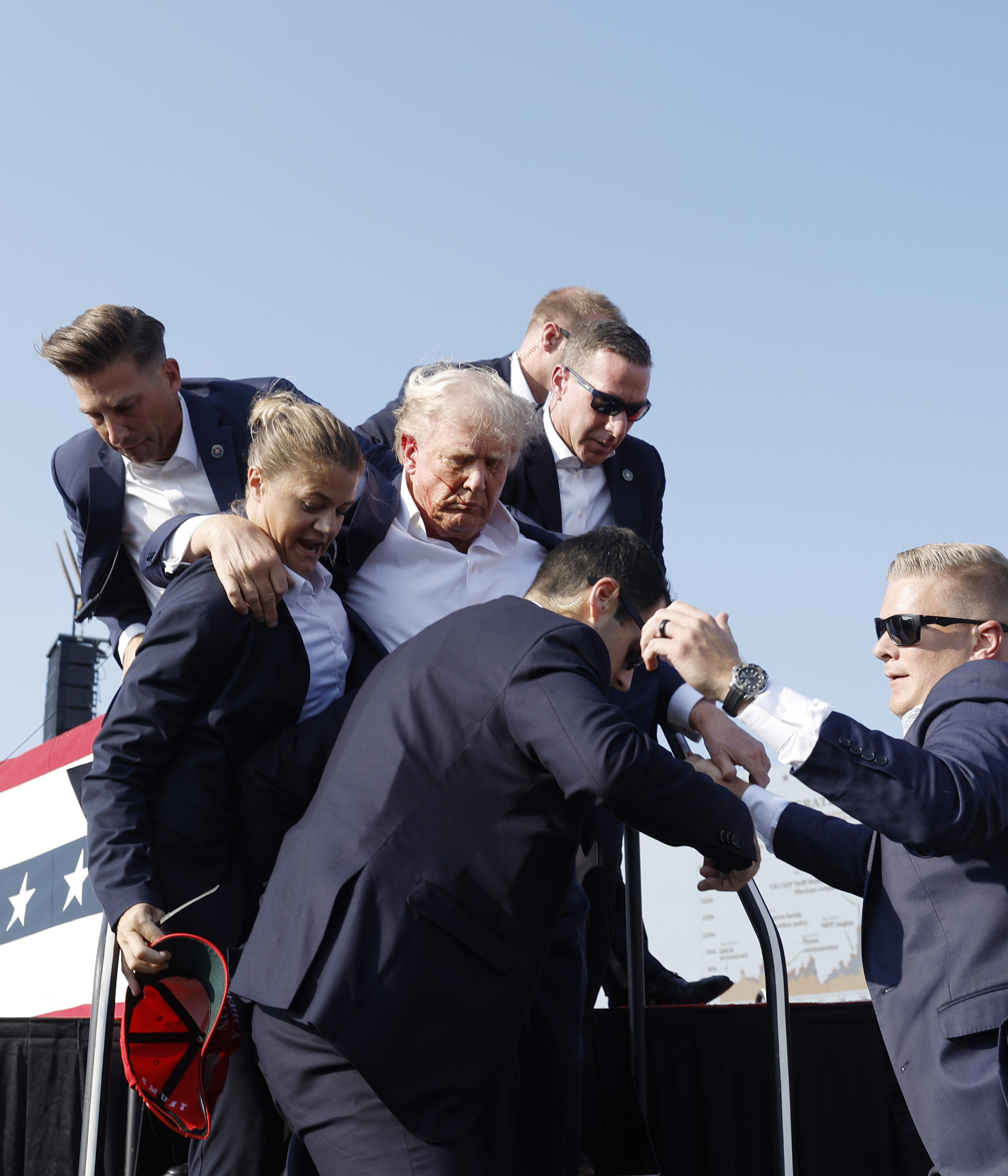 A group of six people, in formal attire and some wearing sunglasses, assist an elderly man in a white shirt down a few steps from a stage decorated with stars and stripes.