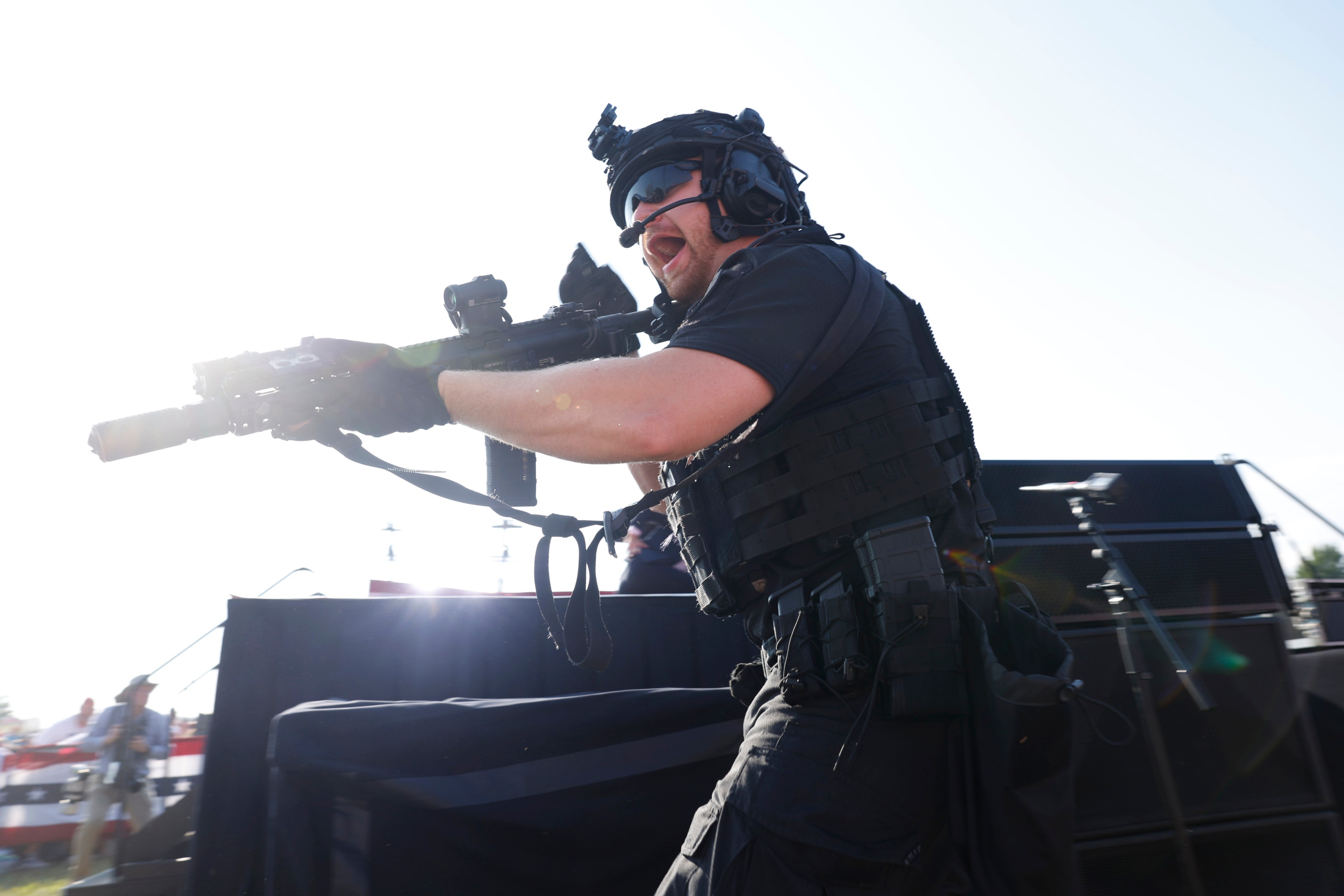 A heavily armed man in tactical gear and helmet, holding a rifle, shouts during daylight in an outdoor setting; background shows onlookers and a stage setup.