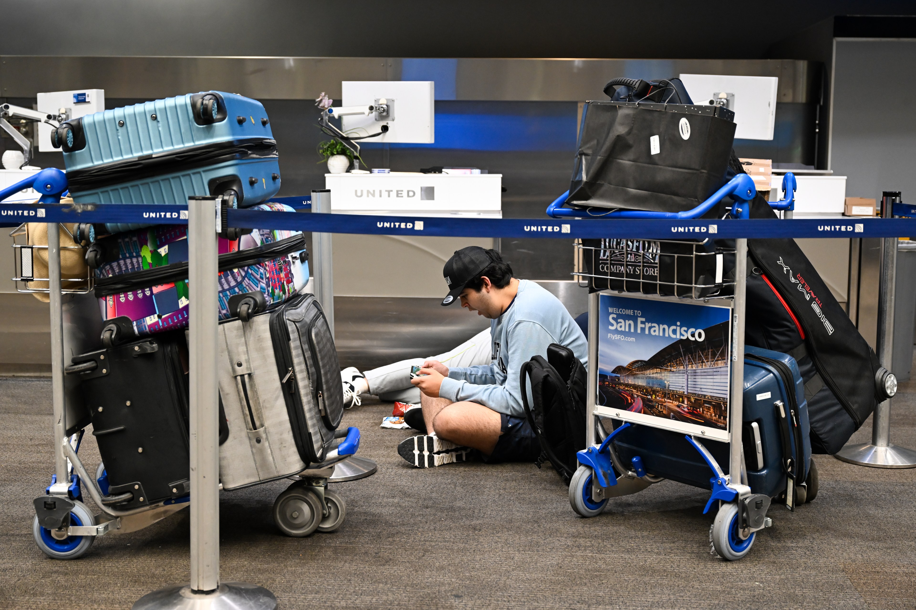 A traveler sits on the floor using a phone, surrounded by multiple luggage carts filled with colorful suitcases at a United Airlines check-in counter in an airport.