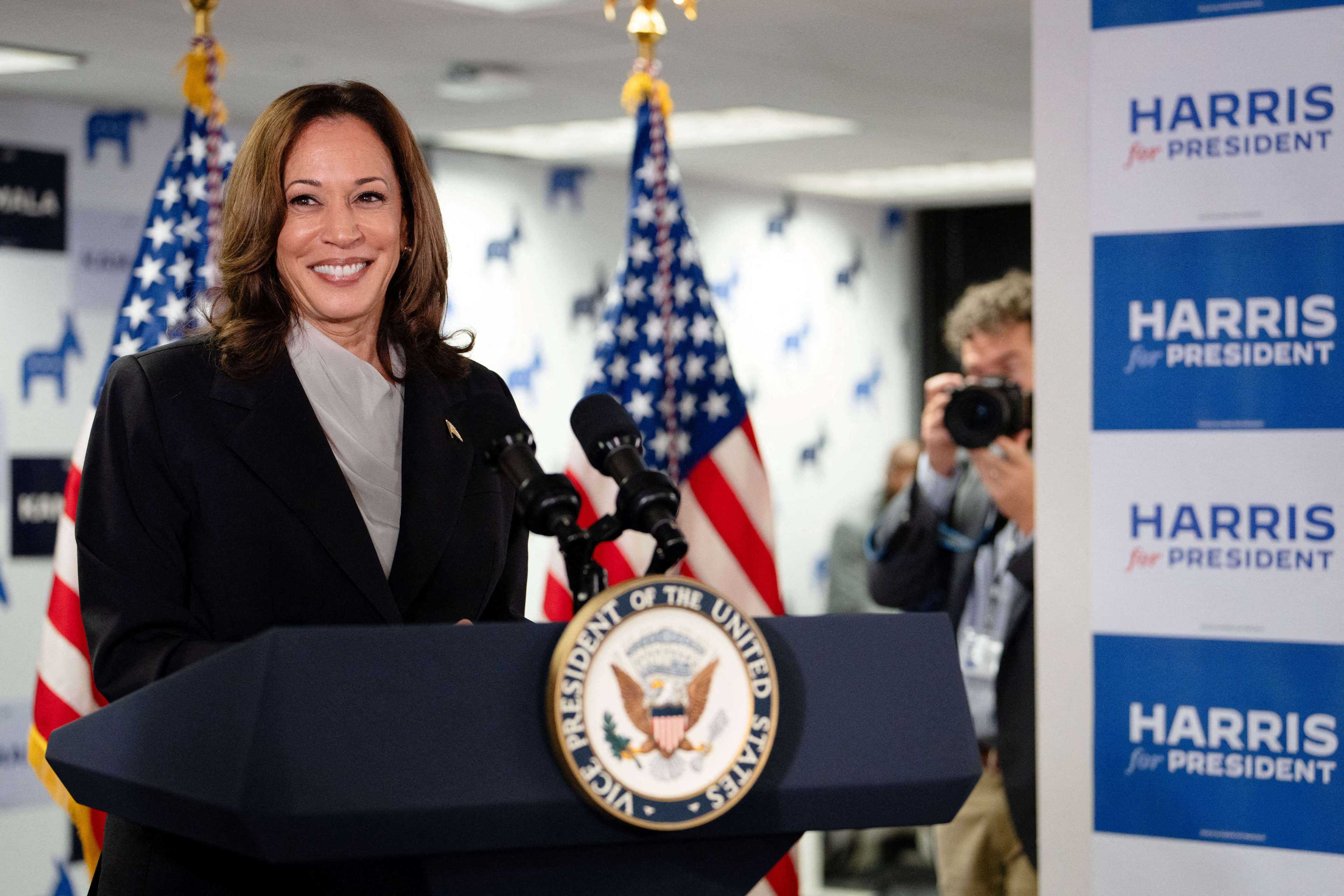 A woman stands at a podium with the U.S. Vice President seal, smiling, surrounded by American flags. Signs reading &quot;Harris for President&quot; are visible in the background.