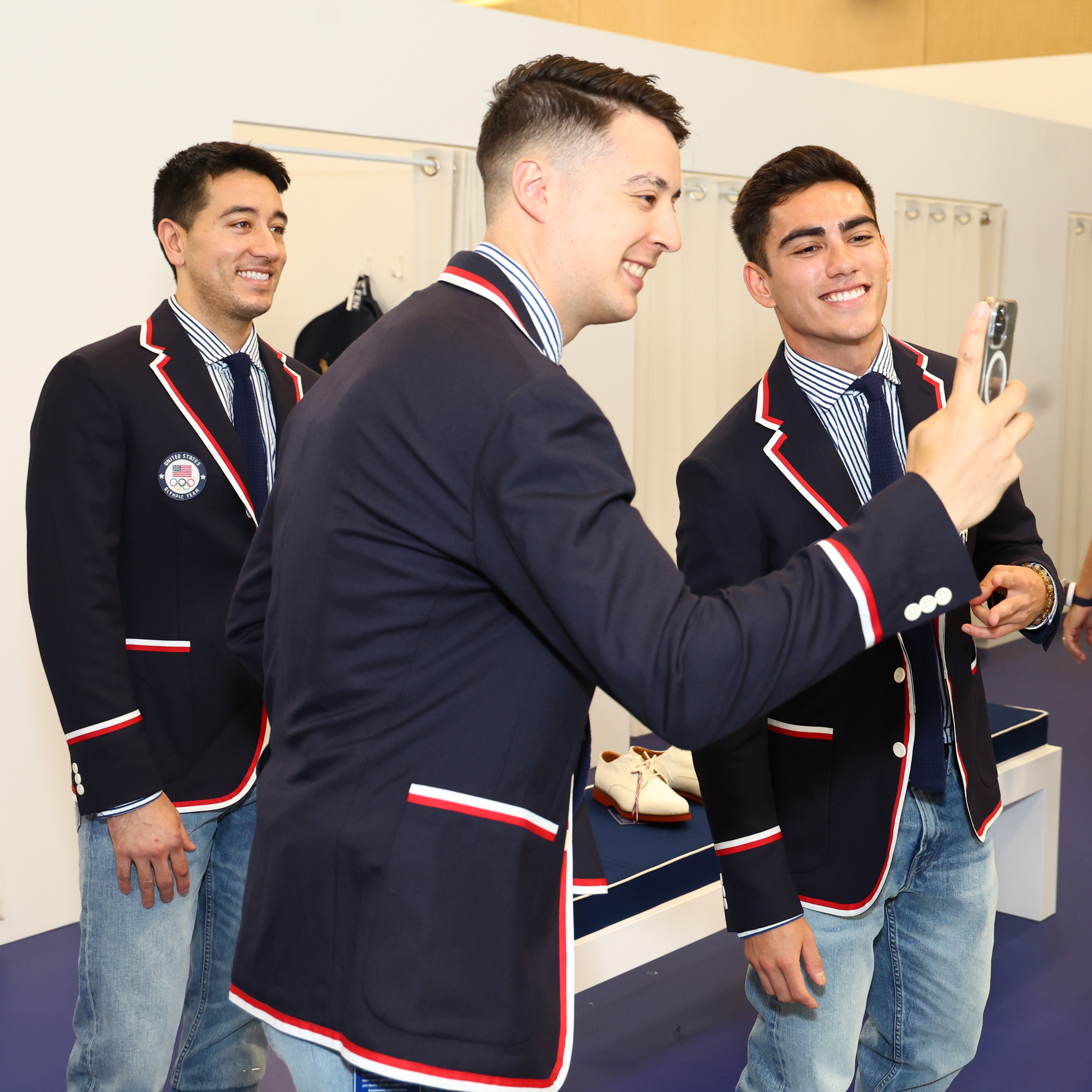 Three men wearing blue blazers with red and white trim and jeans take a selfie. Two other people are seen in the background, standing next to a &quot;Team USA&quot; sign.