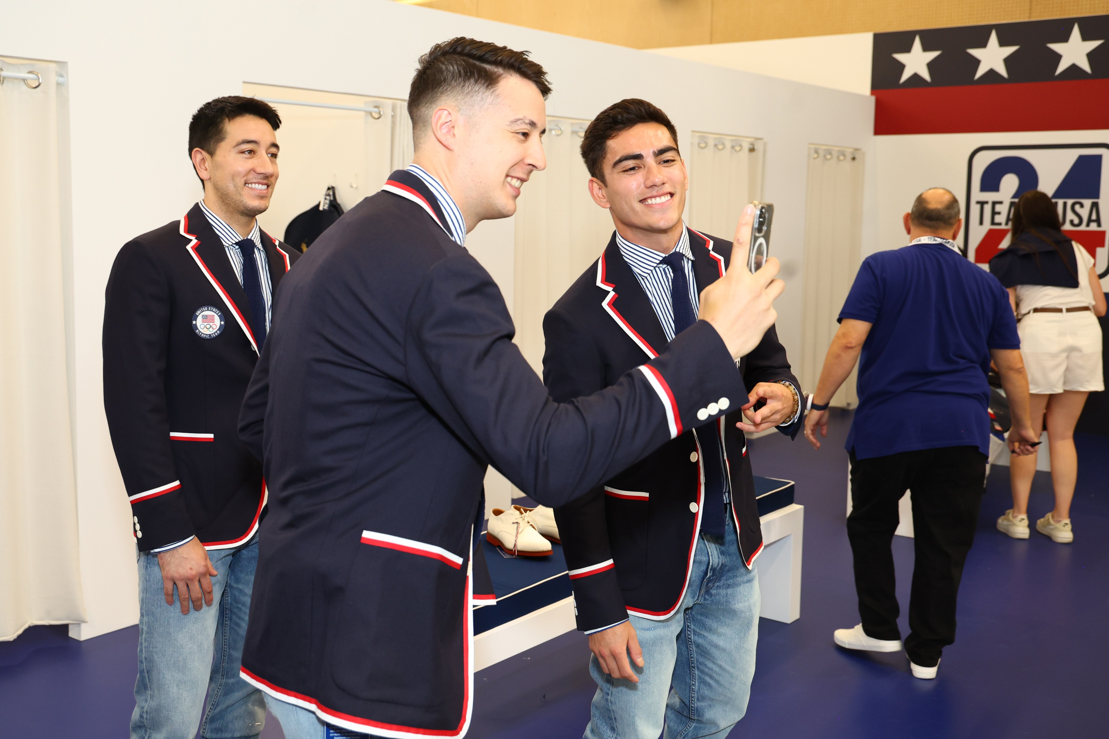 Three men wearing blue blazers with red and white trim and jeans take a selfie. Two other people are seen in the background, standing next to a &quot;Team USA&quot; sign.