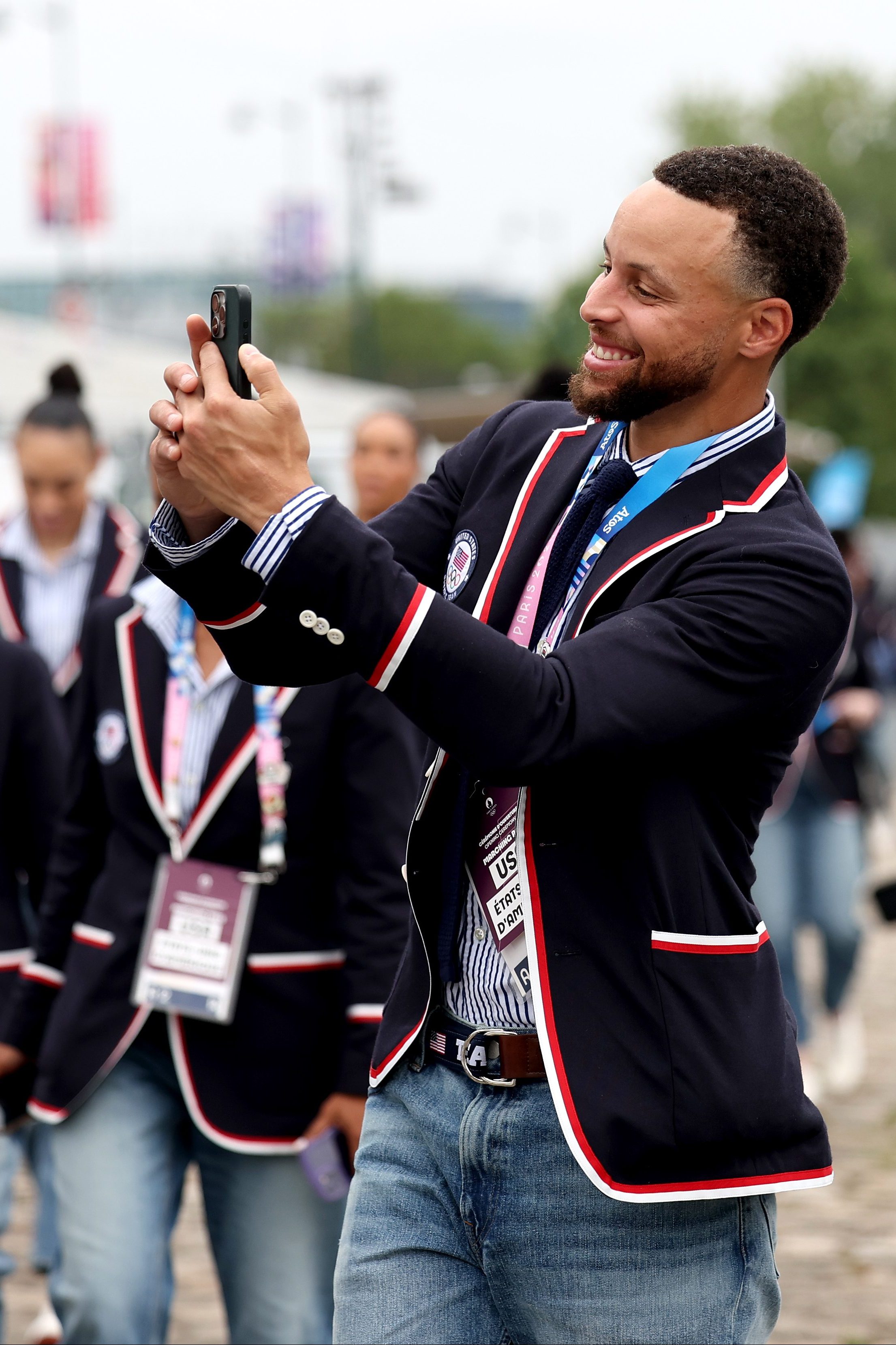 A group of people in matching navy blazers with red and white trim, some wearing blue jeans, are walking outdoors; one man is taking a selfie.