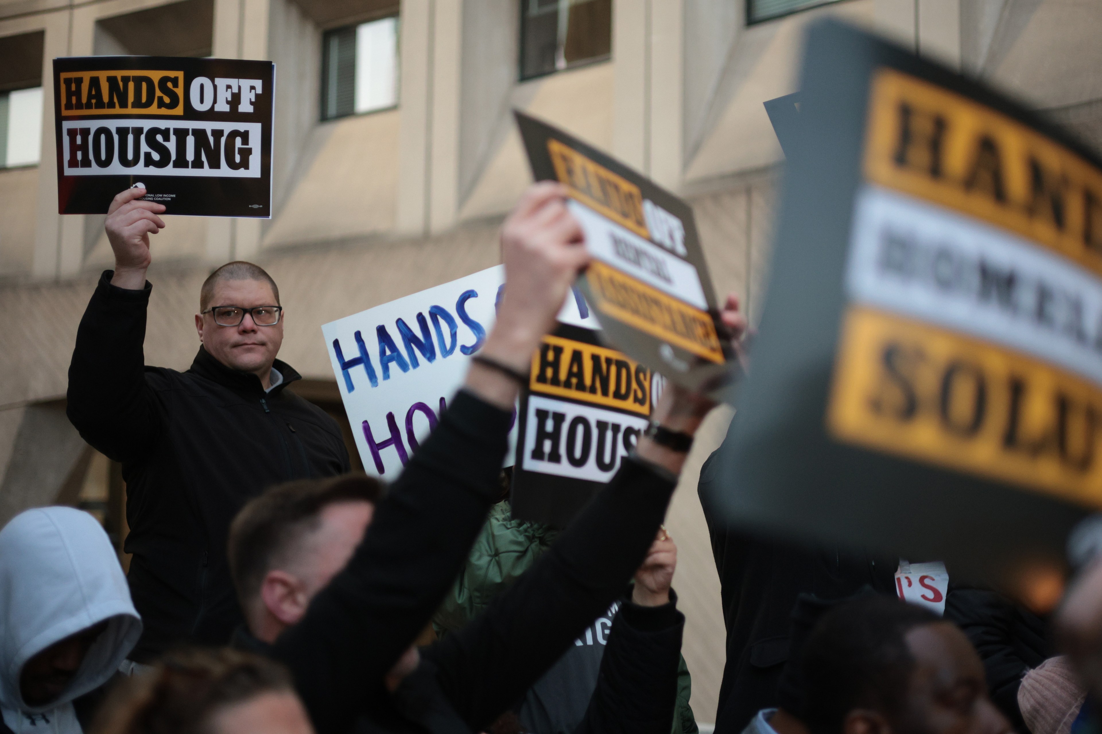 People are gathered holding protest signs that read &quot;Hands Off Housing.&quot; They stand in front of a building, expressing a unified message.