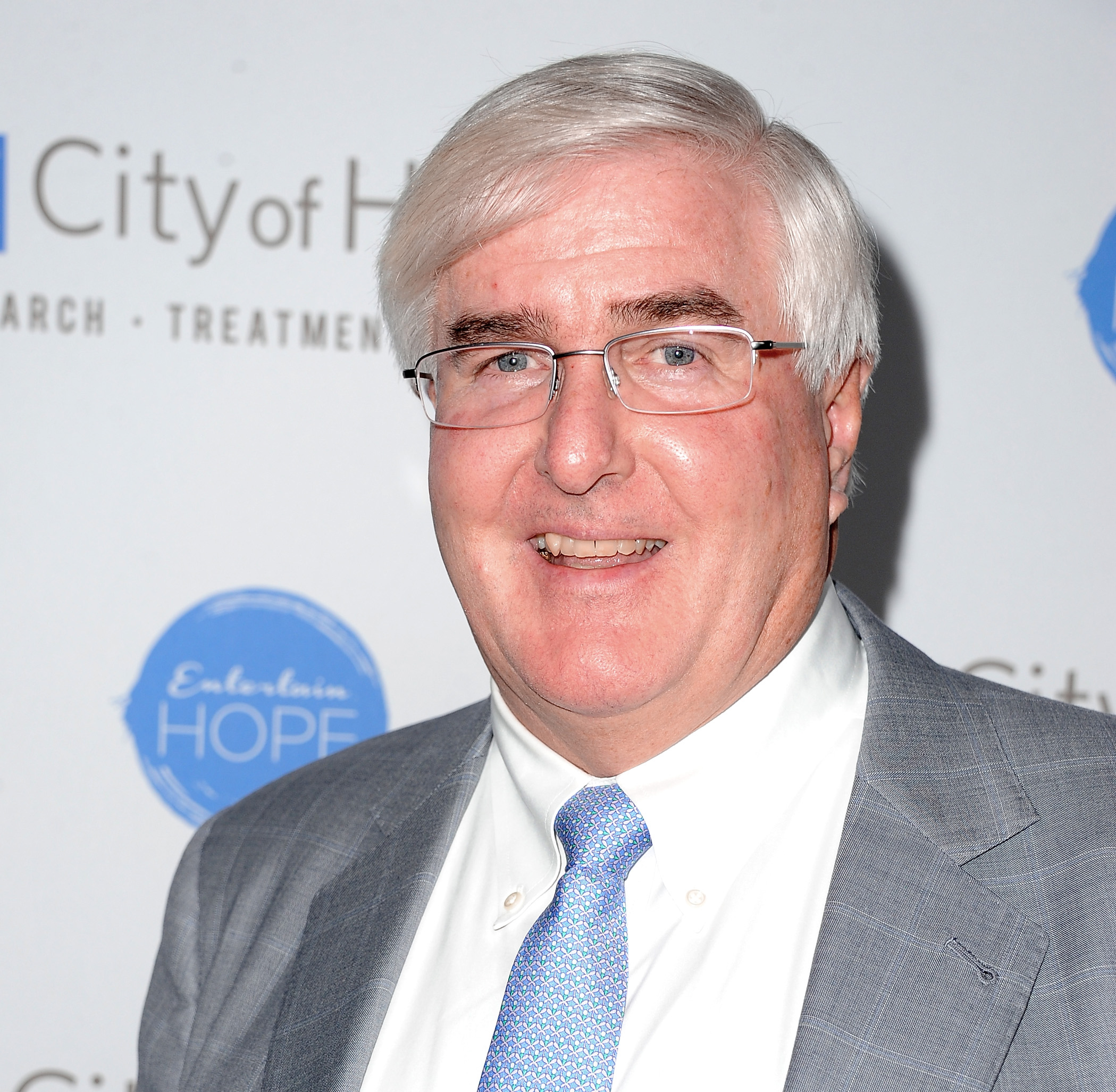 A man with short, white hair, glasses, and a grey suit with a light blue tie smiles at the camera. The backdrop has the text &quot;City of Hope&quot; and a blue logo.