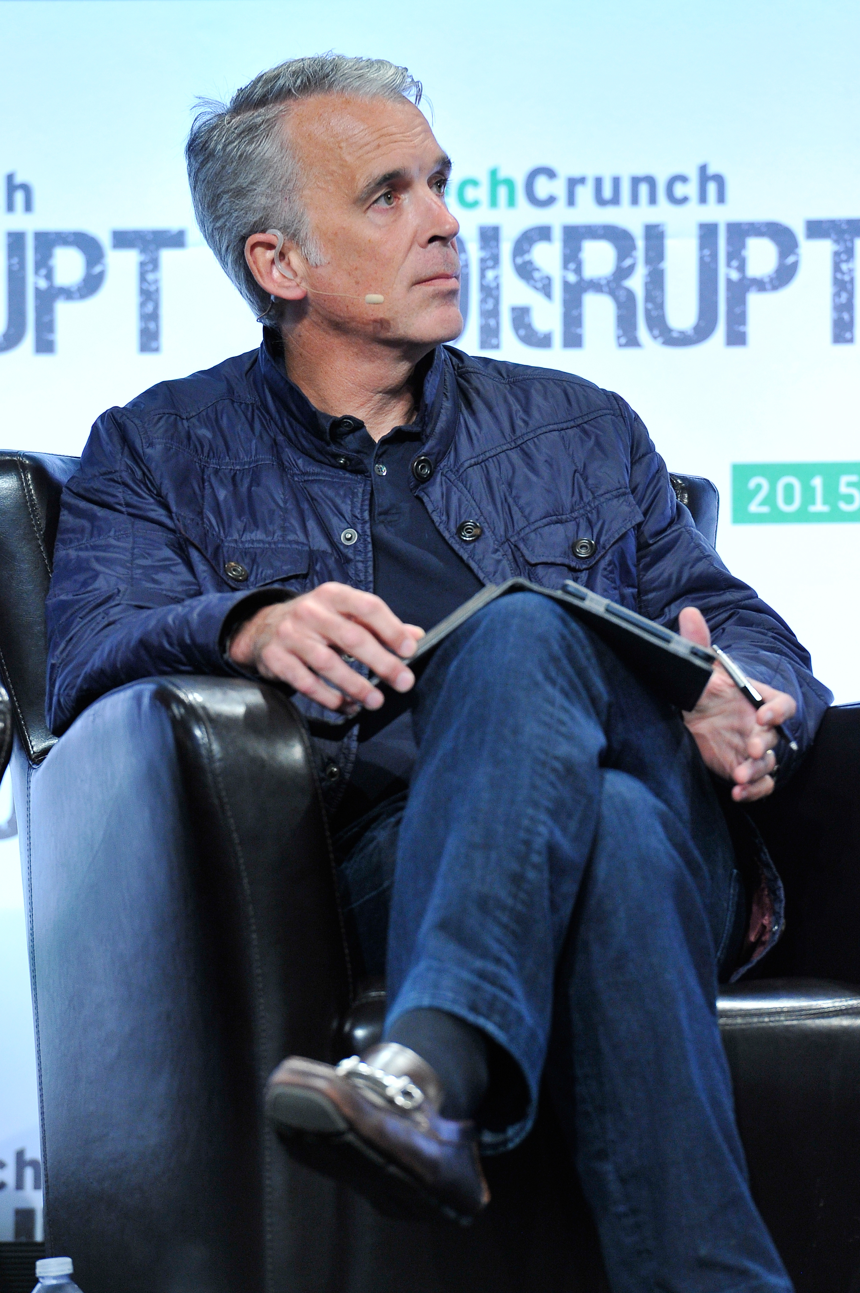 A man with short gray hair sits on a black chair, holding a tablet. He wears a dark blue jacket, jeans, and brown loafers. The background reads "TechCrunch Disrupt 2015."