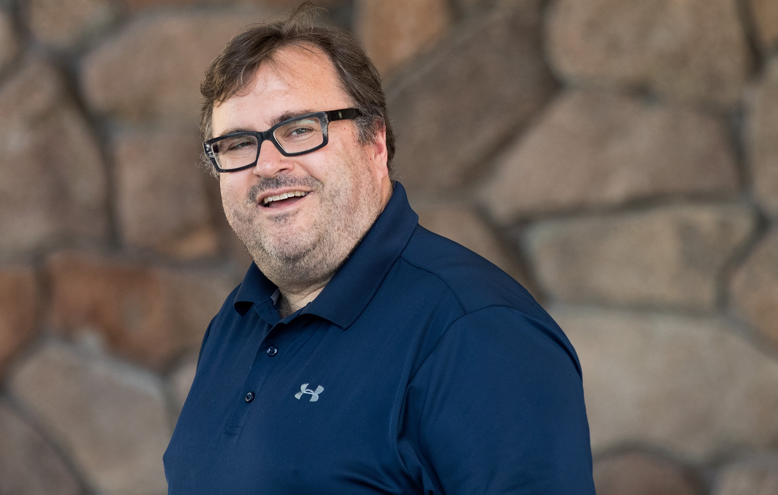 A smiling man with glasses and messy hair is wearing a navy blue Under Armour polo shirt. He stands in front of a stone wall background.