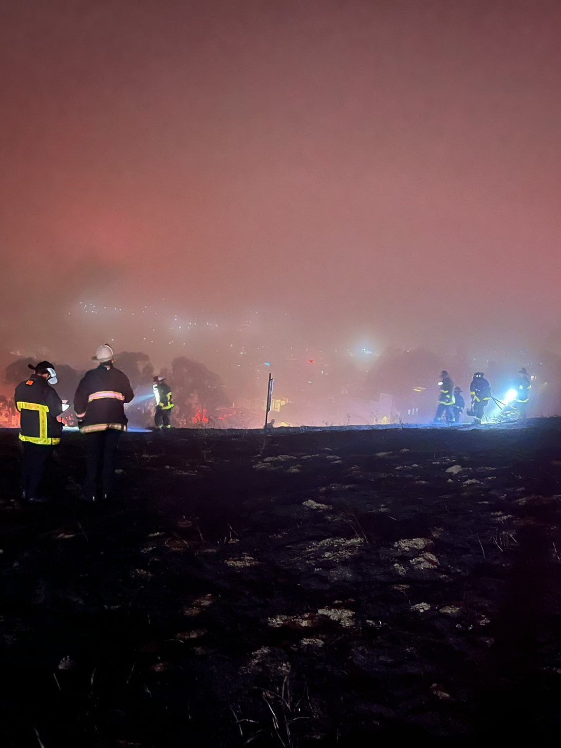 Under heavy nighttime fog, several firefighters use flashlights and tools to check a grassy park area burned by fire using flashlights and tools for hot spots.