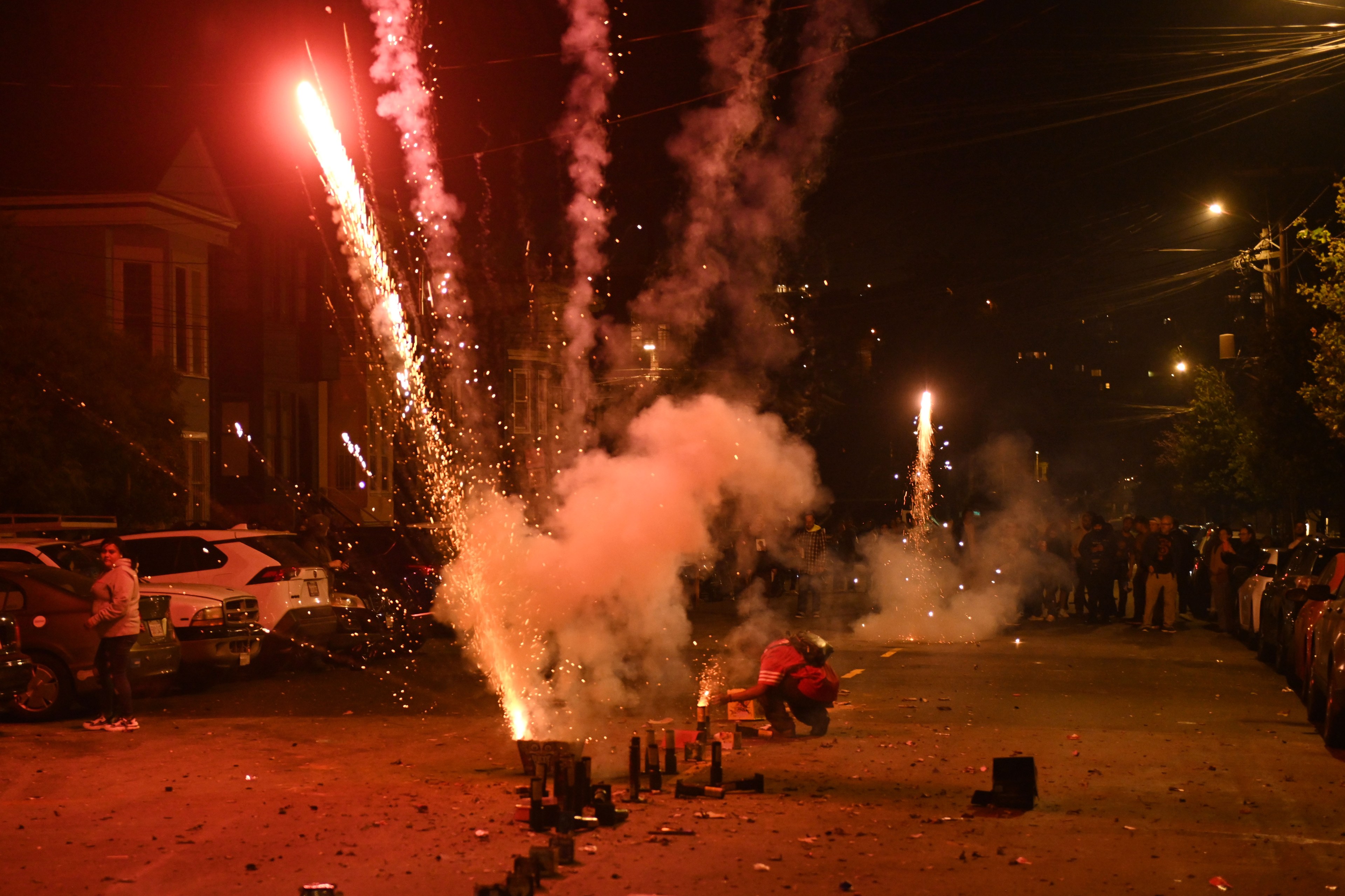 At night on a street, fireworks are being launched, with sparks and smoke filling the scene. People are gathered on either side, some watching or tending to the fireworks.