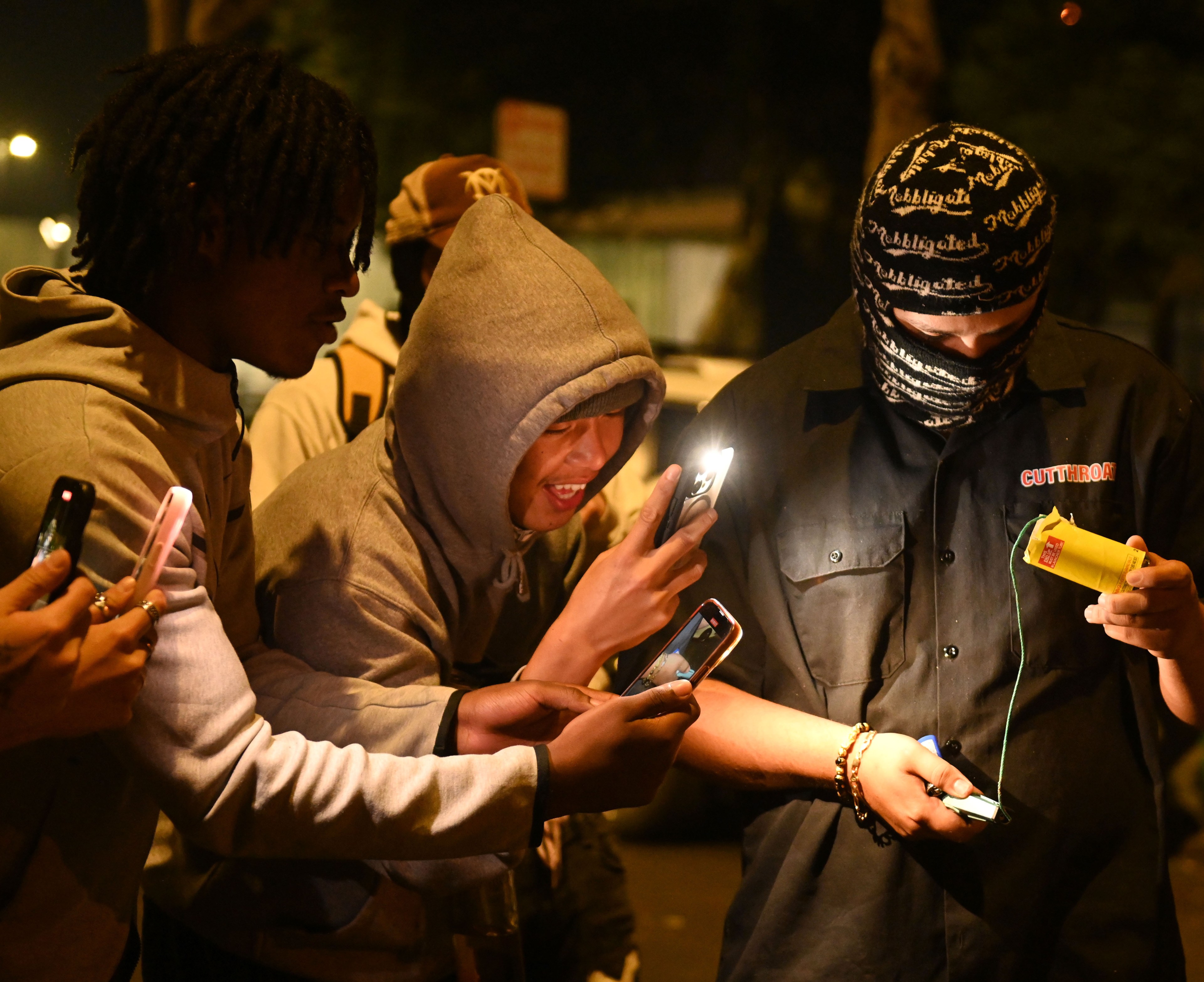 Several people gather at night, using smartphones to illuminate a man in a dark shirt and mask, who is holding a cylindrical object with a fuse.