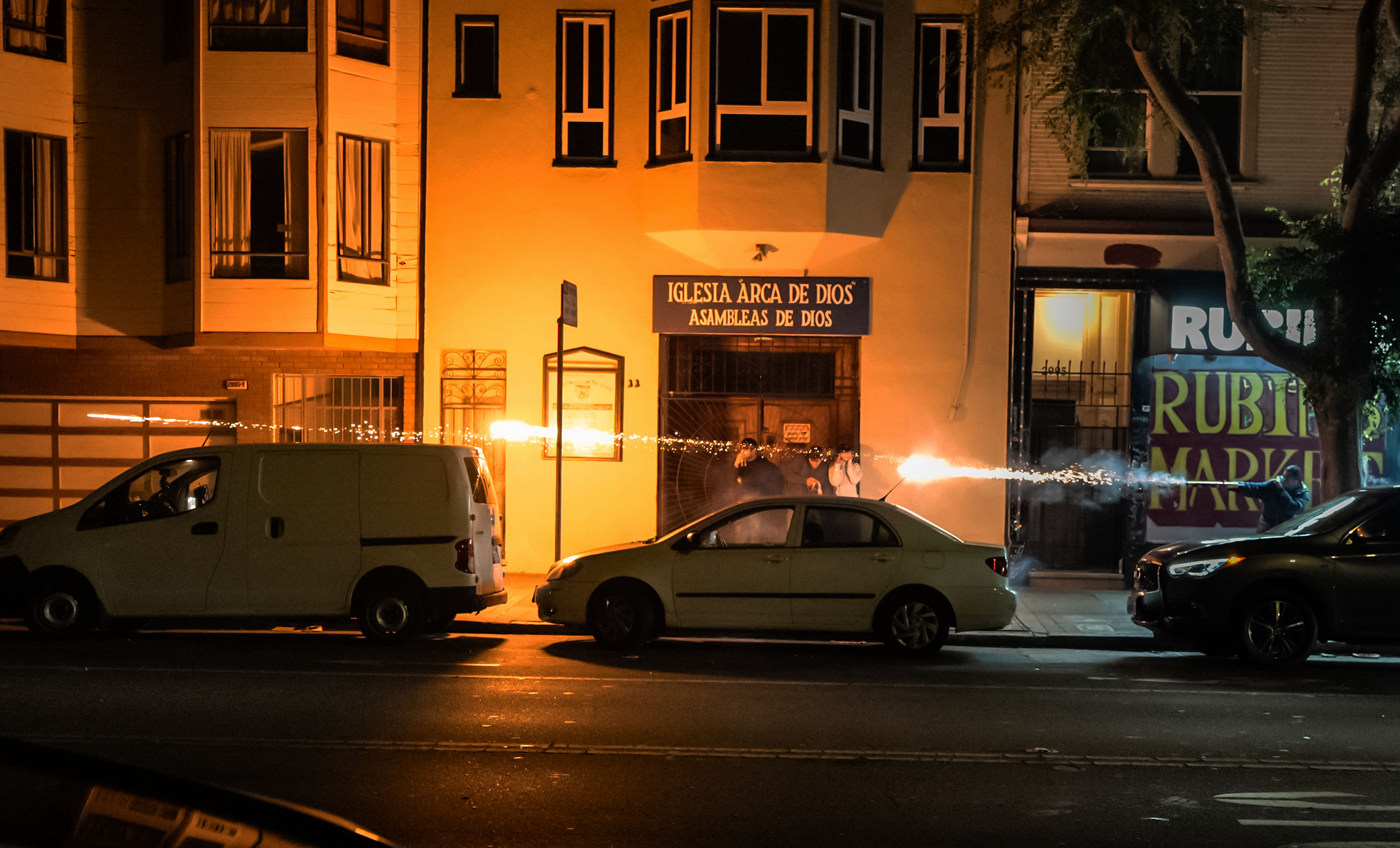 Several people, centered around a church entrance, discharge fireworks among parked vehicles on a dimly lit street at night. Sparks light up their paths.