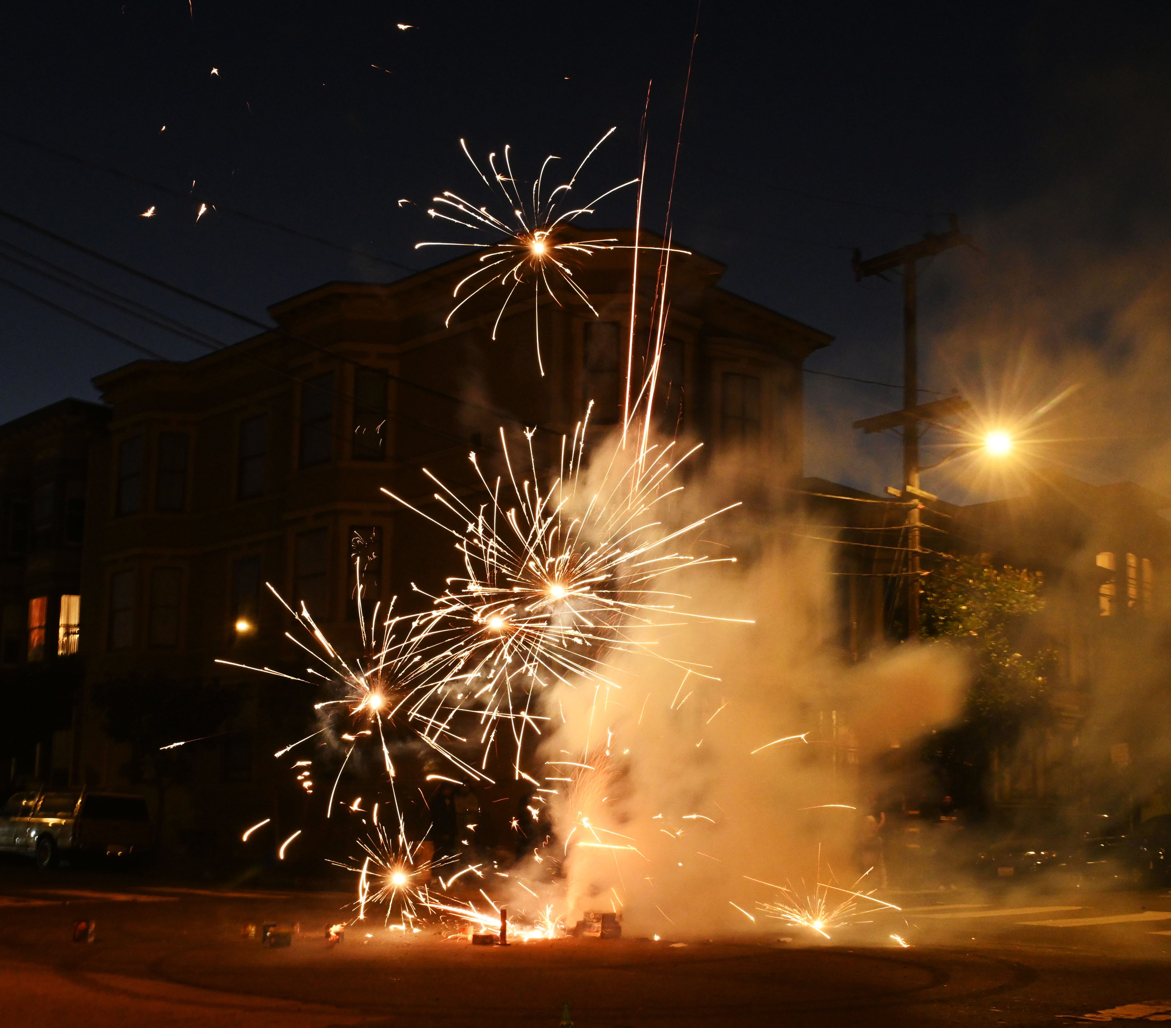 In a dimly lit street at night, fireworks burst and spark, lighting up the scene with bright trails and smoke in front of darkened buildings.