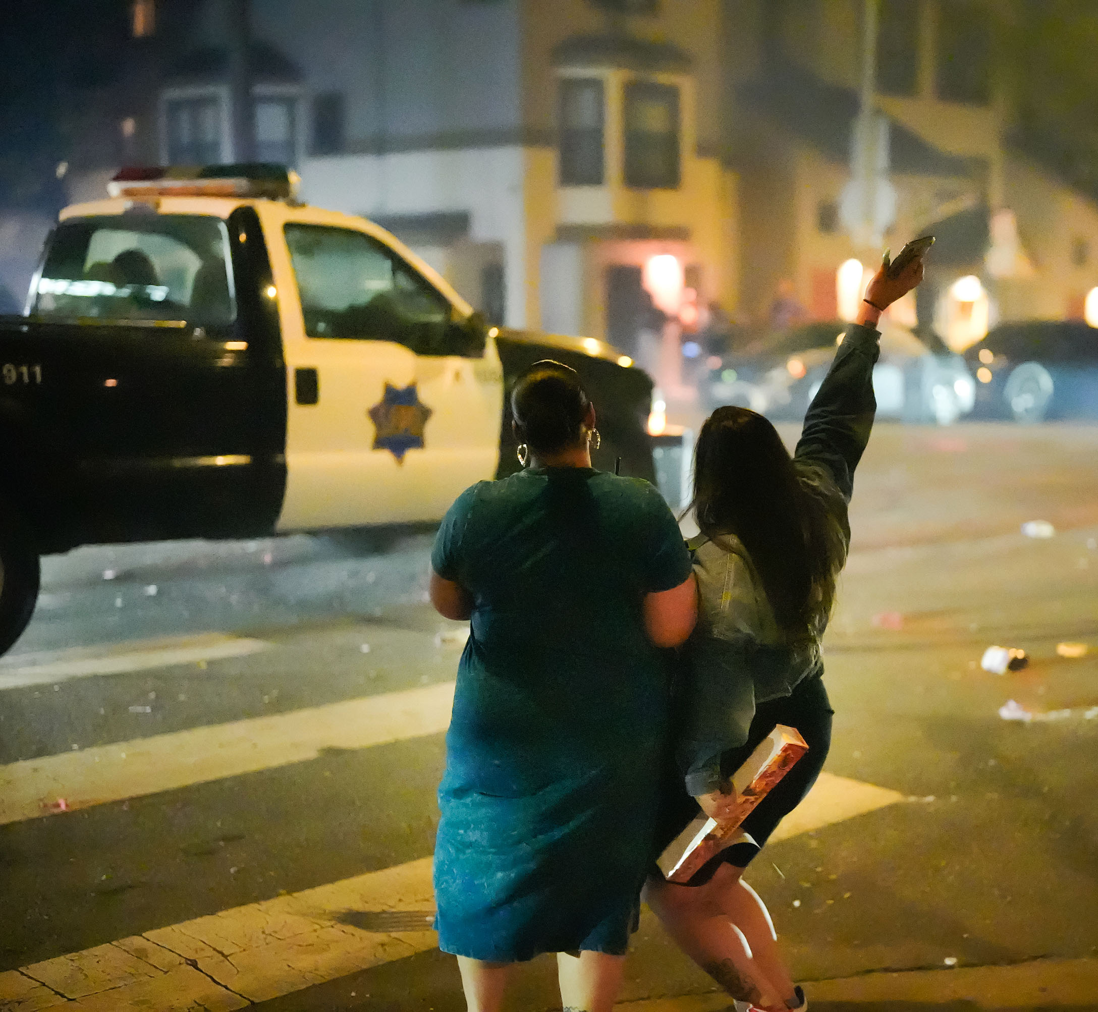 Two people are on a street corner at night, facing a police vehicle. One waves an object in the air while the other stands beside them holding a box.