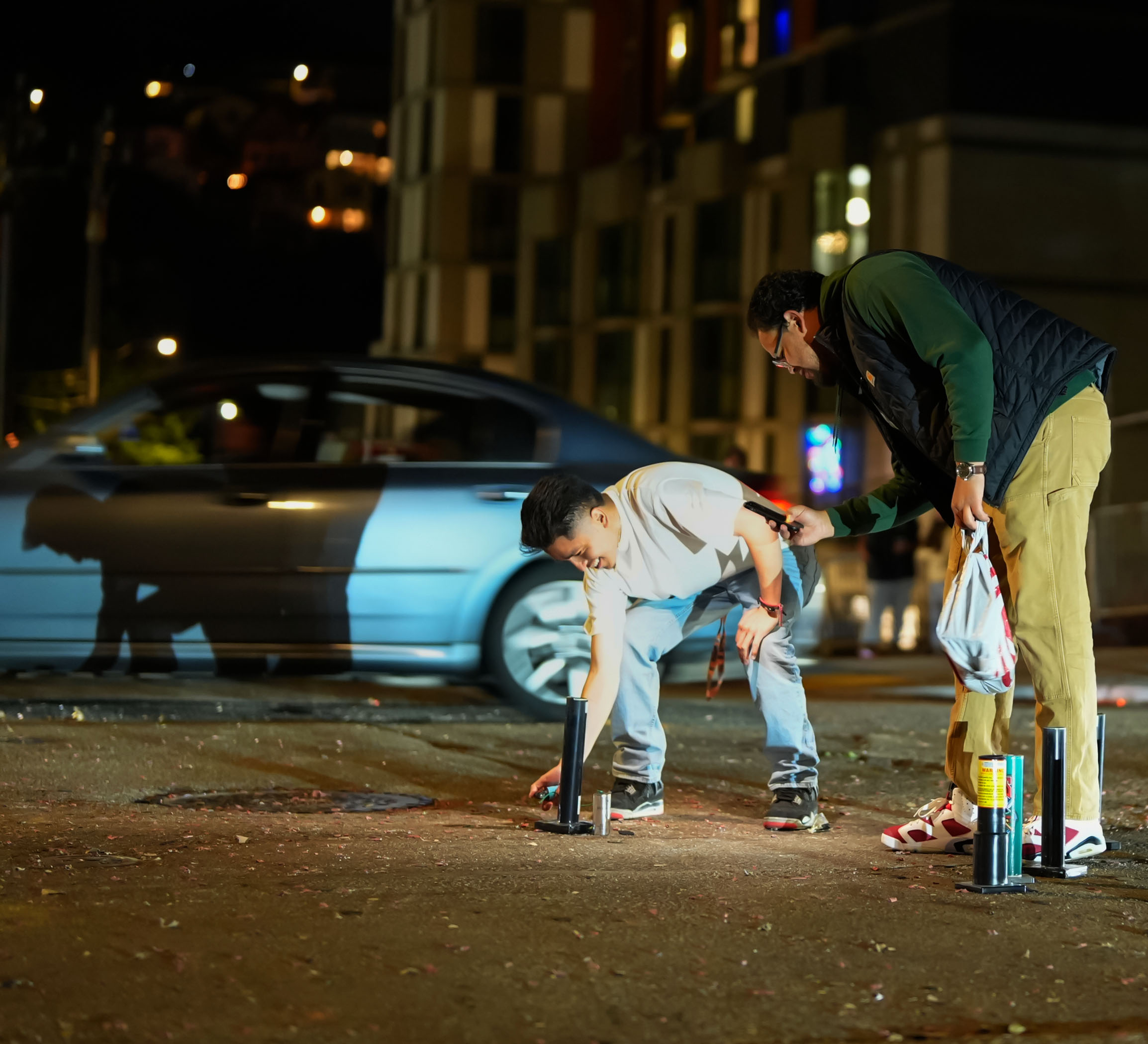 Two people are setting up fireworks on the street at night. One person is crouching down placing a firework tube, while the other holds a flashlight and some bags.