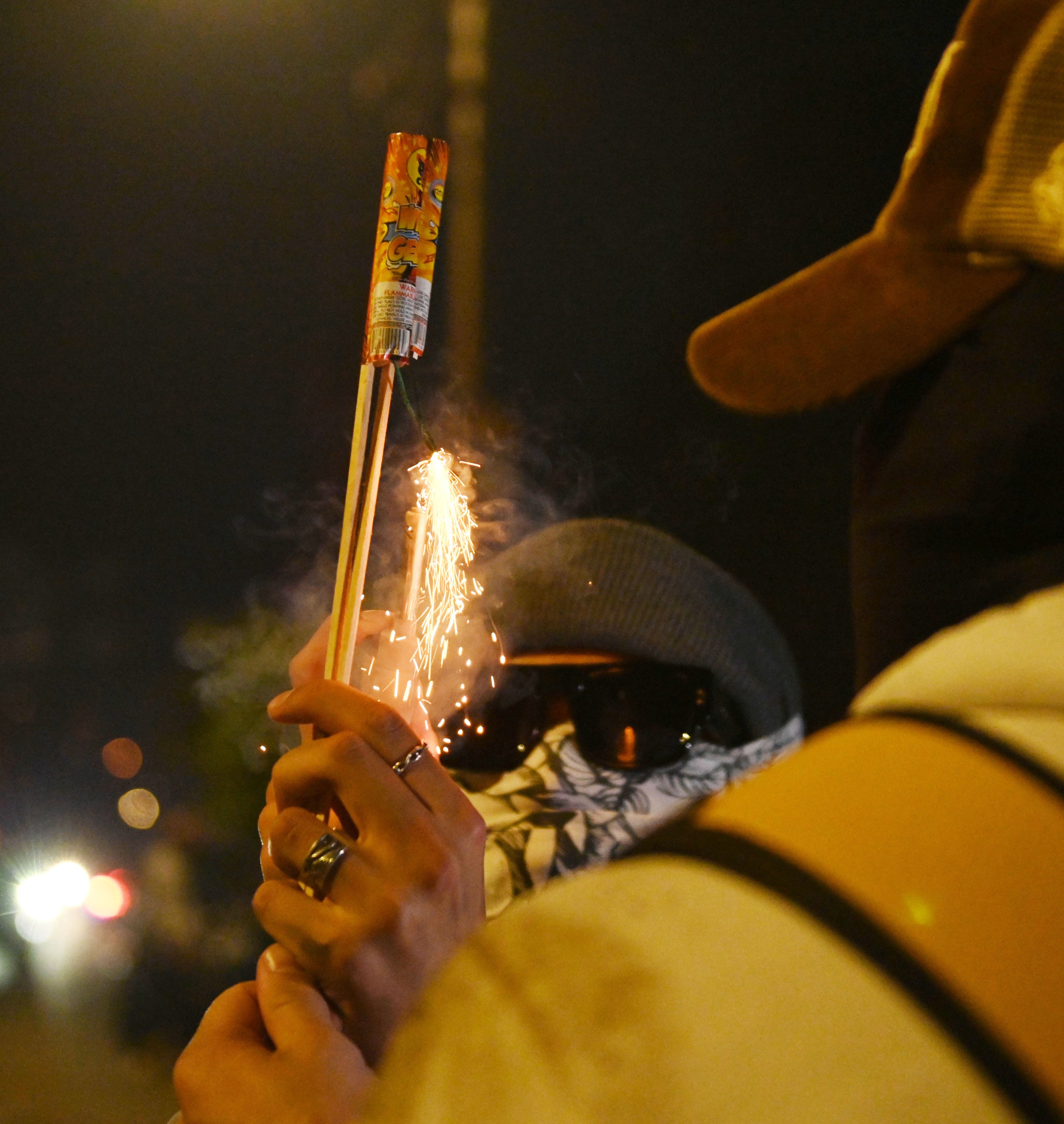 A person in a beanie and mask is lighting a firecracker, which is sparking, at night.