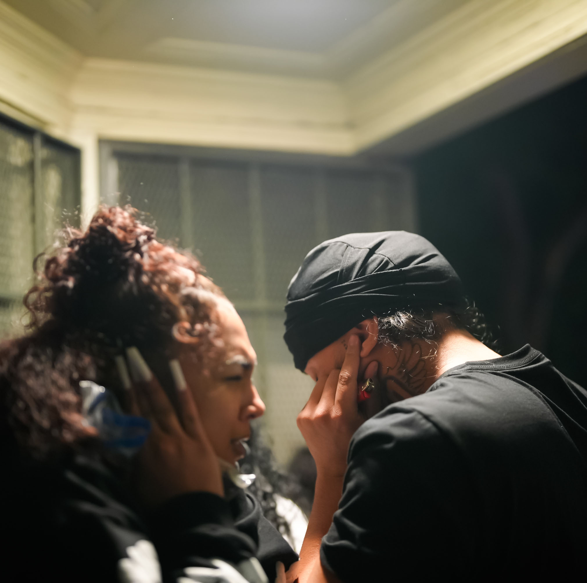 In a dimly lit scene, two people are standing close together, covering their ears. One has curly hair tied up and the other wears a black headwrap.