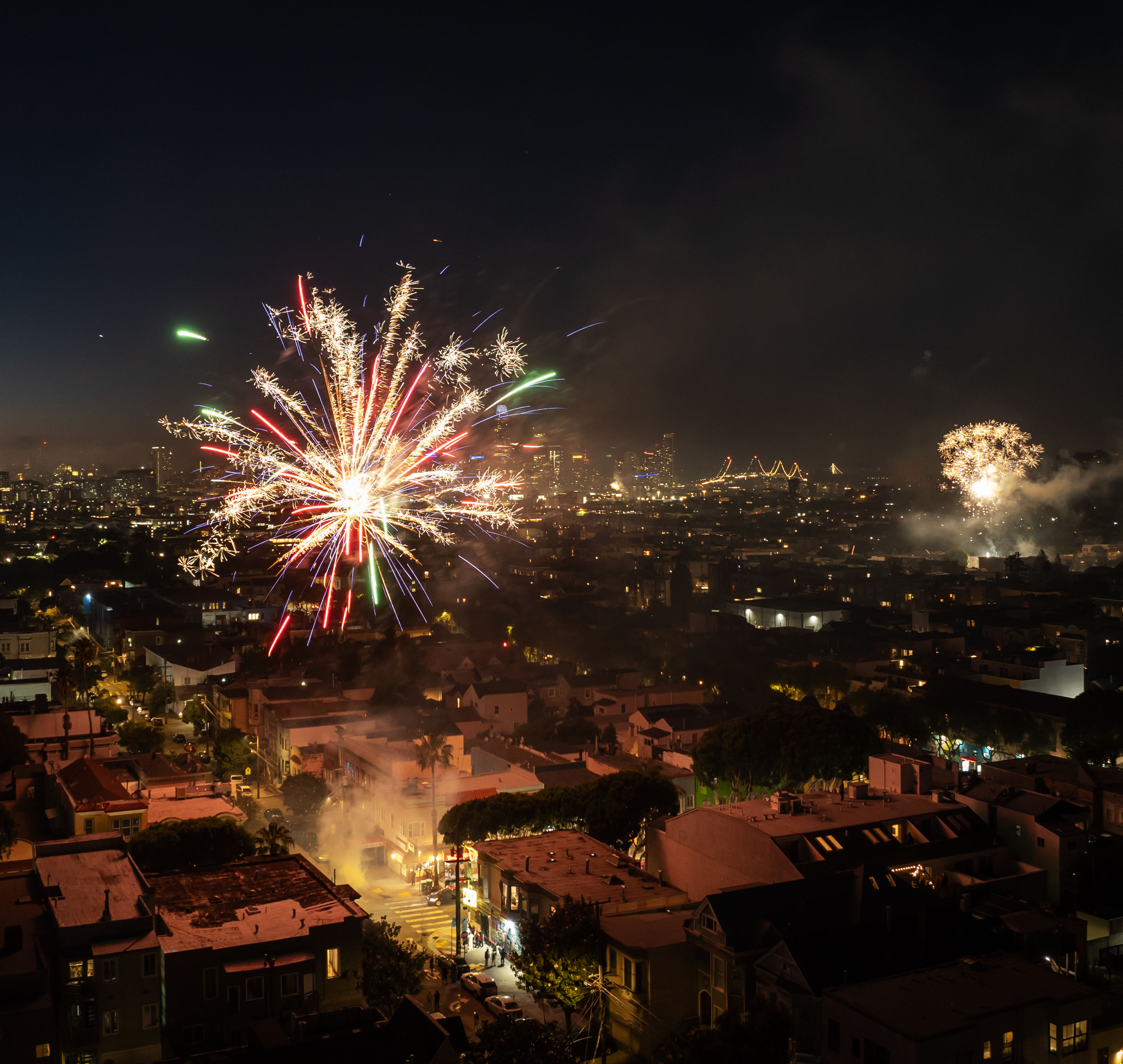 The image shows a nighttime cityscape with vibrant fireworks. Houses and streets are illuminated in the foreground, while a bustling city with bridges is in the background.