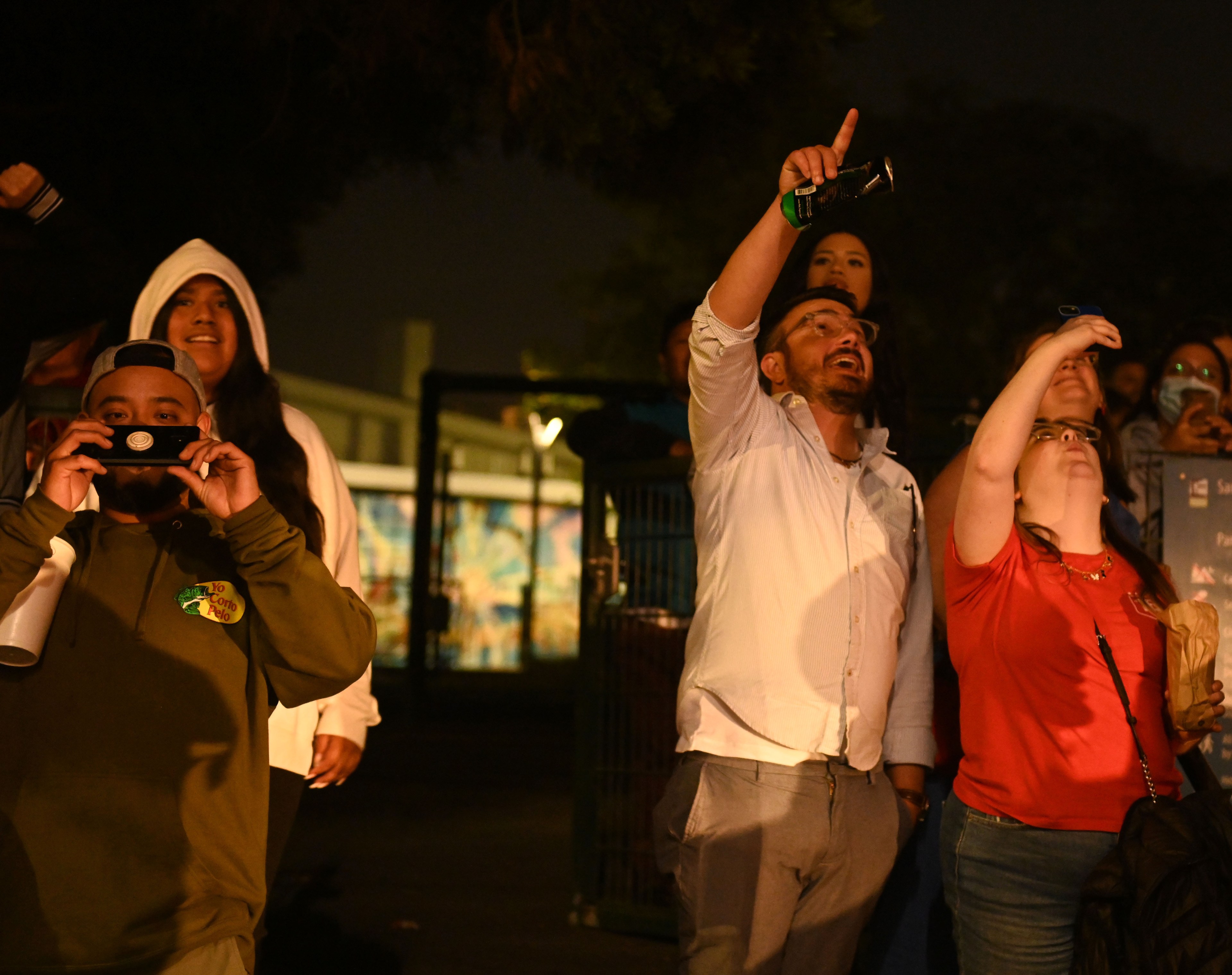 A group of people stands outside at night, looking towards fireworks in the sky. One person in a hoodie holds a phone, possibly taking a picture, while others seem engaged and curious.