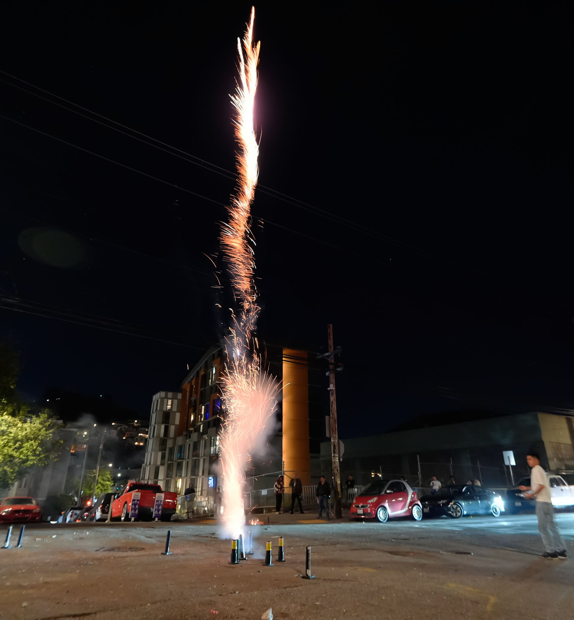 A firework shoots up, sparkling against the night sky in an urban area, people and cars are nearby, with illuminated buildings in the background.