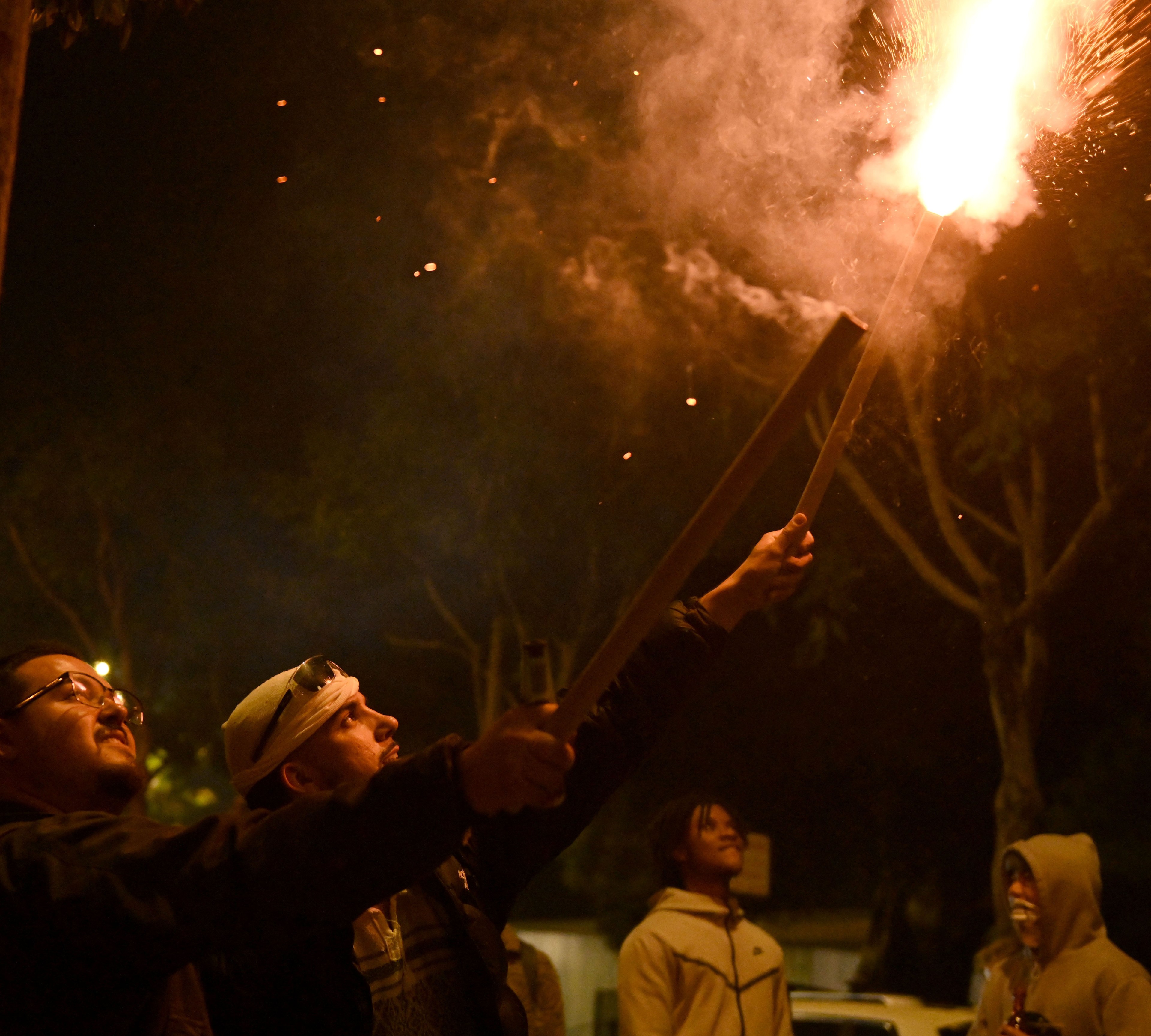 Two people hold large, lit fireworks sticks, creating a bright flare and smoke against the night sky. Others watch in the background, with trees and soft streetlights visible.