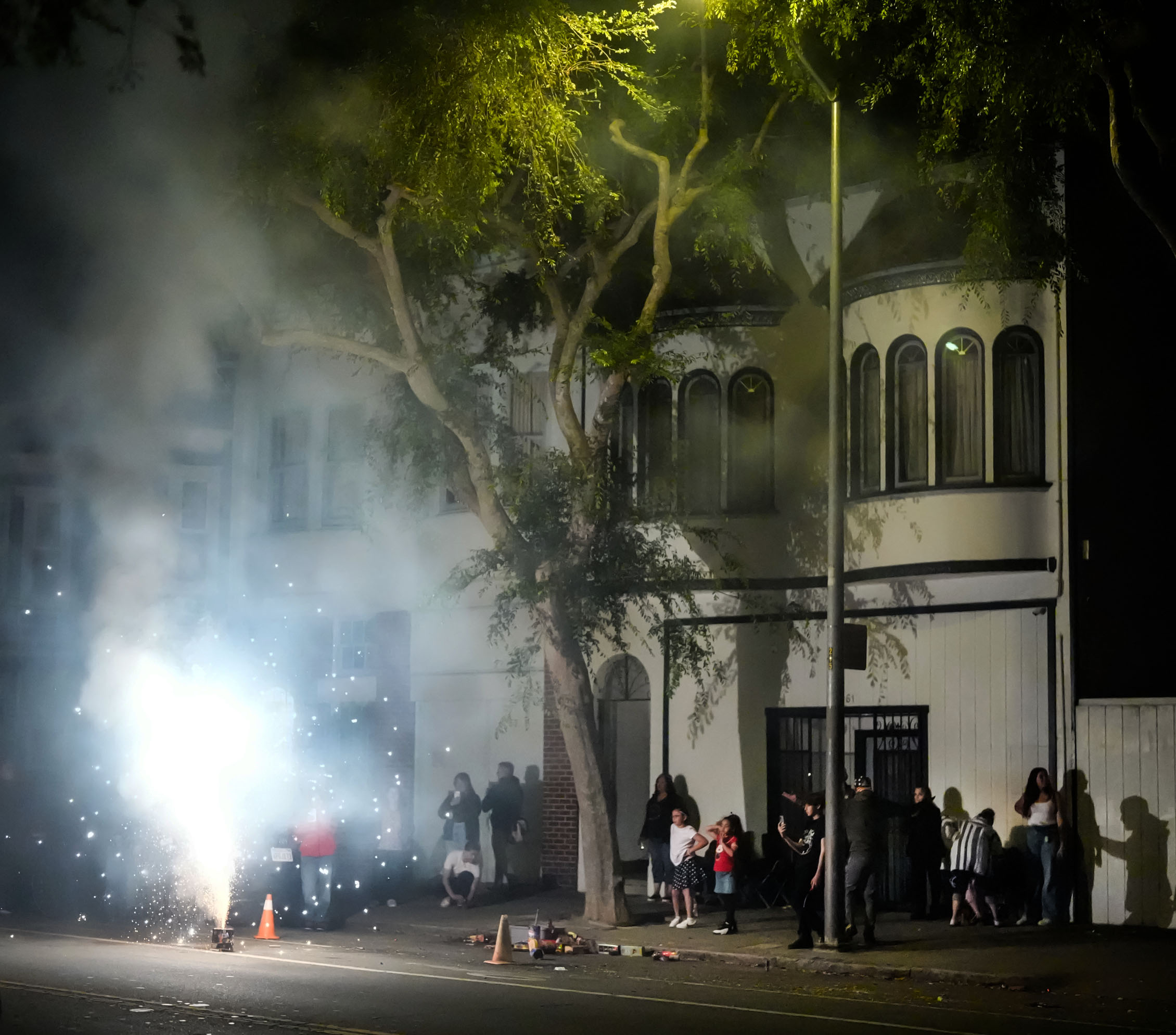 At night, a group of people gather outside a building while a bright flash and smoke from fireworks illuminate the scene; cones and debris lie on the street.