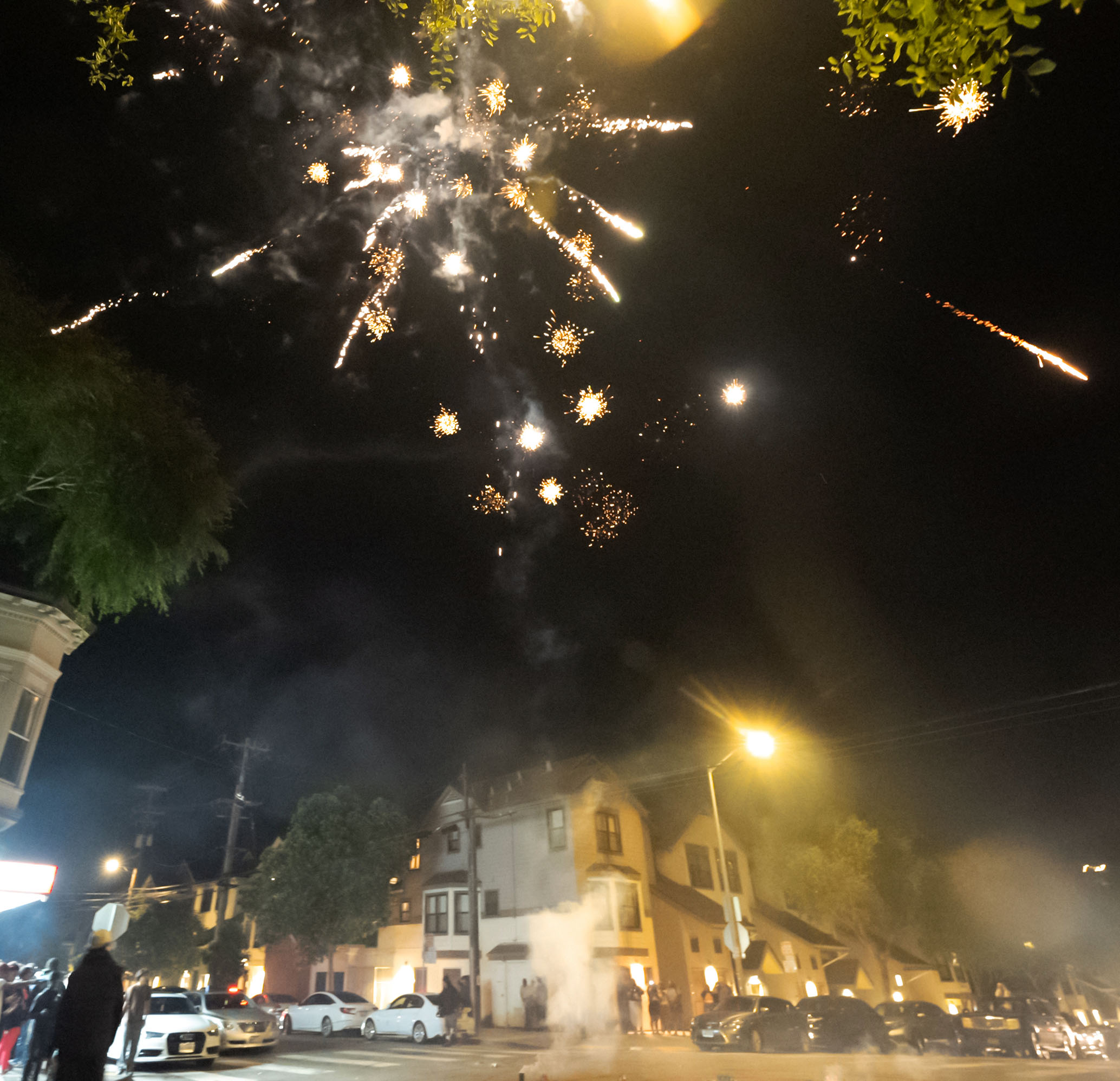 The image shows fireworks exploding in the night sky above a street with houses, cars parked along the road, and people watching the display.