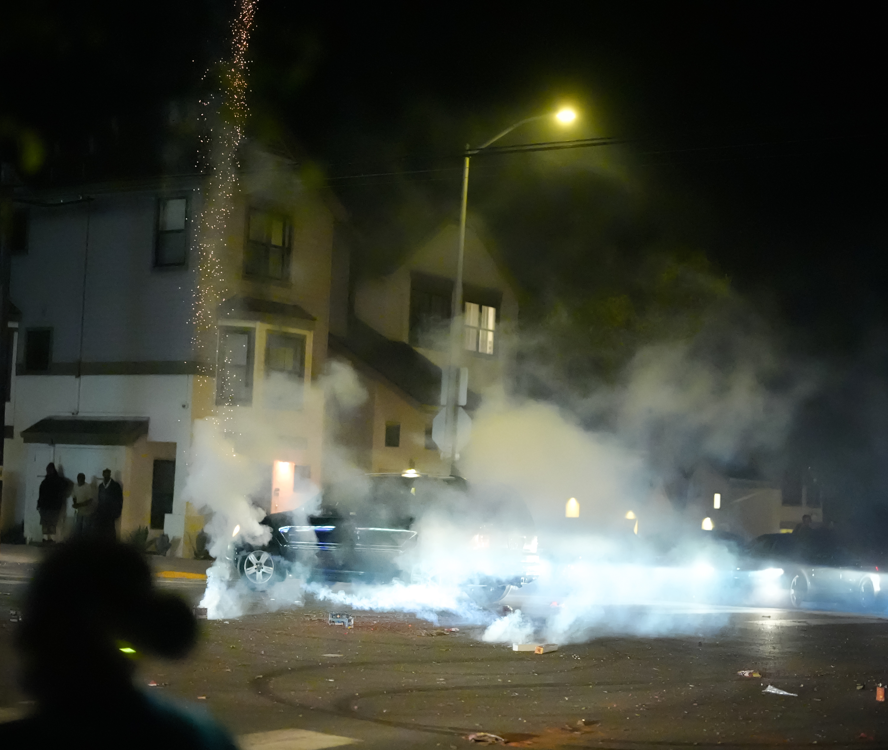 A nighttime scene shows a car driving through thick smoke in an intersection illuminated by a streetlamp. Fireworks or sparks are seen shooting upwards, while people stand nearby.