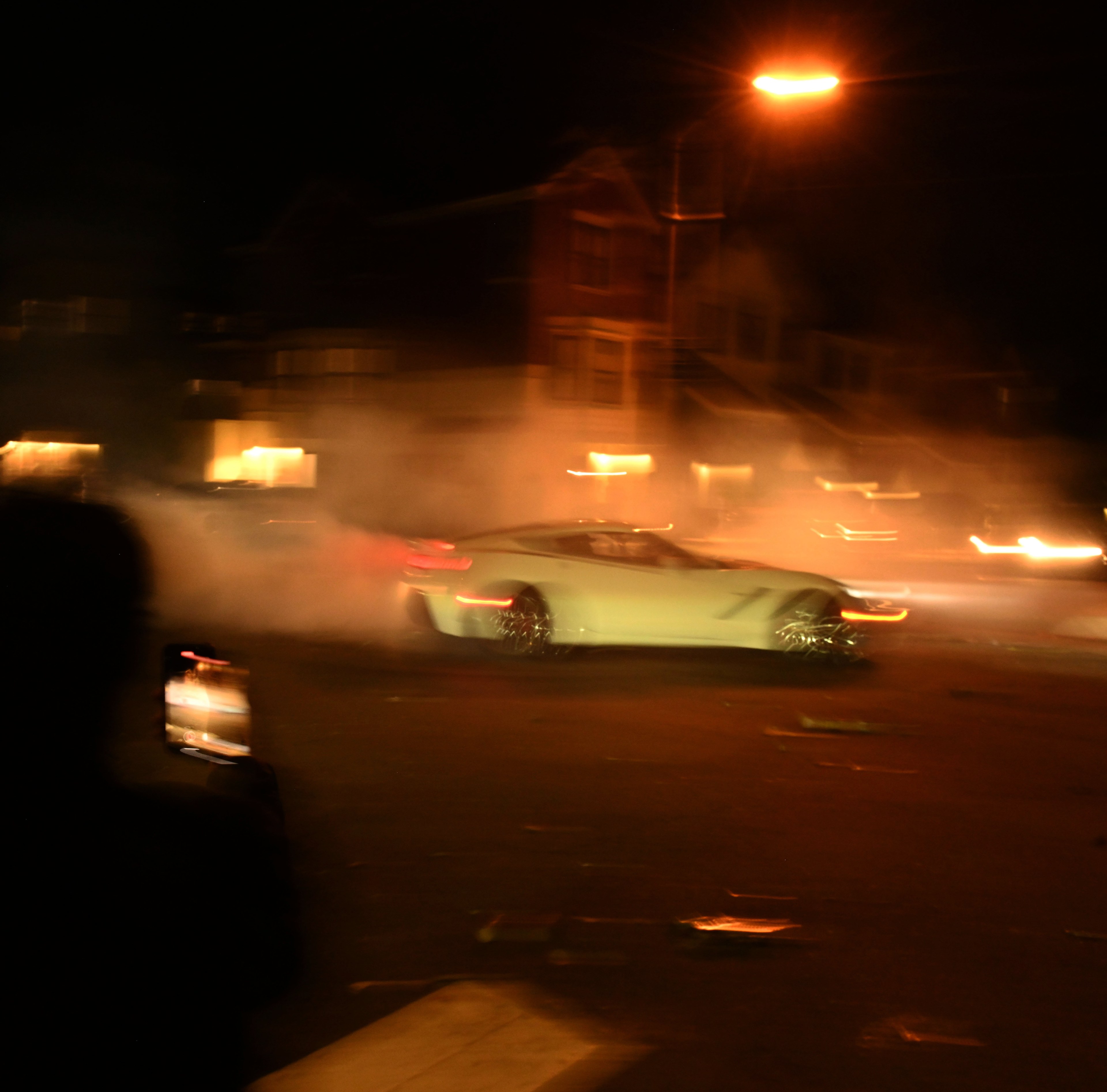 A white sports car is drifting during a sideshow at night, creating a cloud of smoke. Blurry figures with illuminated phones watch and record the event from the street.