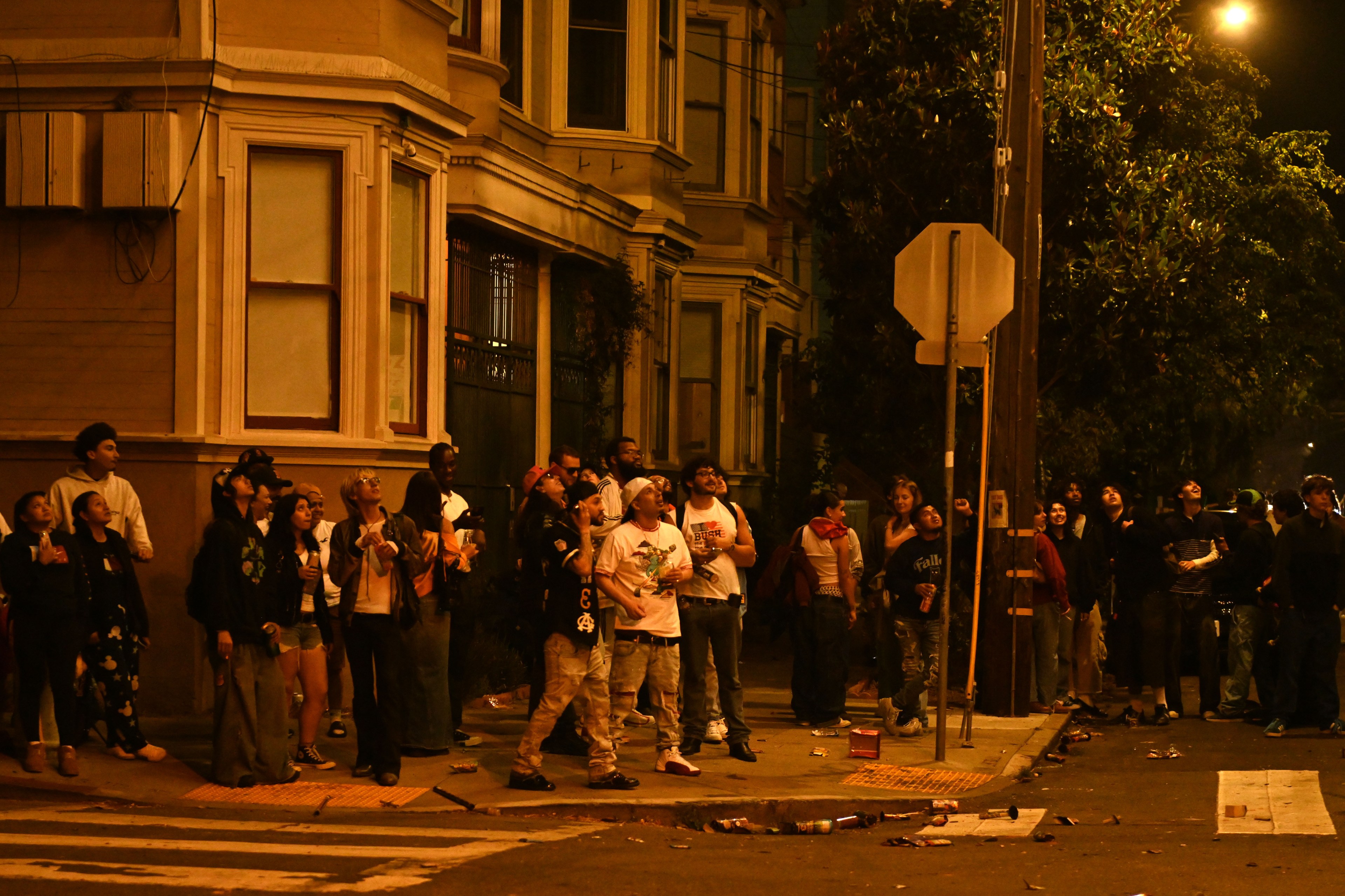 A group of people stand on a city street at night, looking upwards at fireworks in the night sky. They are gathered near a building with bay windows, and the ground is littered with trash.