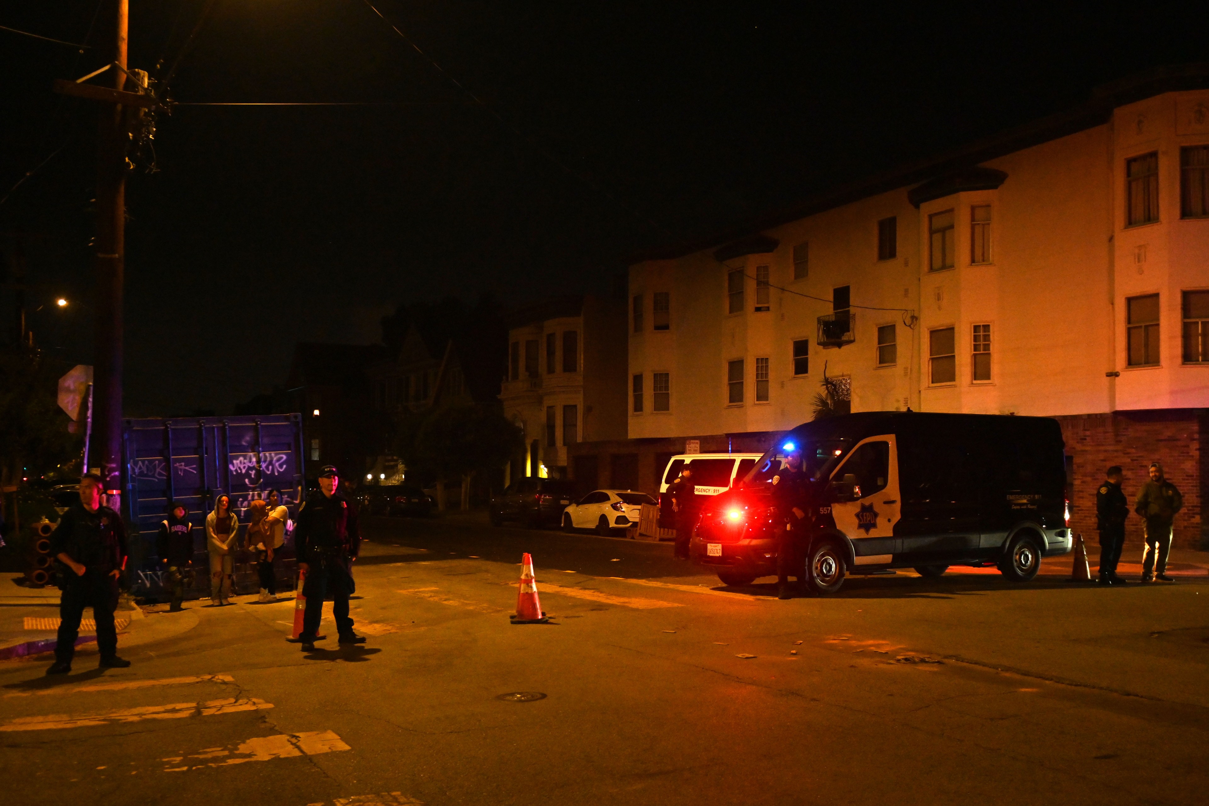 The image depicts a nighttime street scene with multiple police officers and vehicles, orange traffic cones, and a group of people near a building.