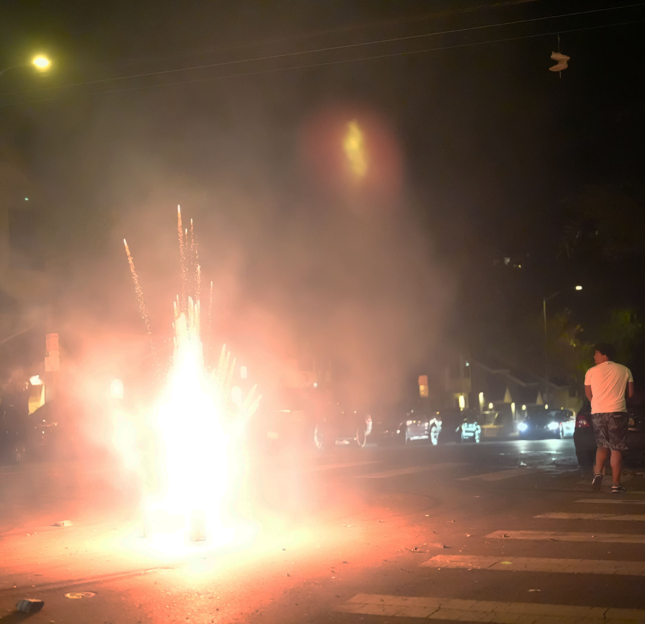 A bright firework display ignites in the middle of a street at night, with sparks and smoke rising from it. A person stands nearby watching the spectacle.