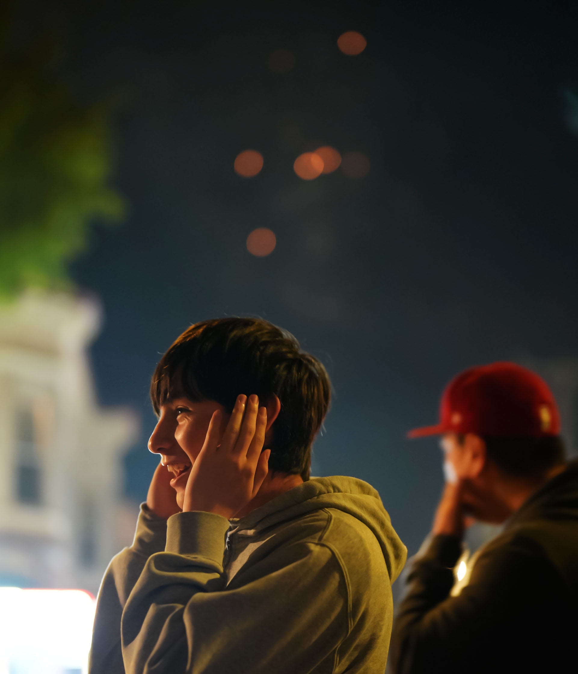 Two people are covering their ears while watching fireworks at night; the person in front is smiling and wearing a hoodie, and the person in the background is wearing a red cap.