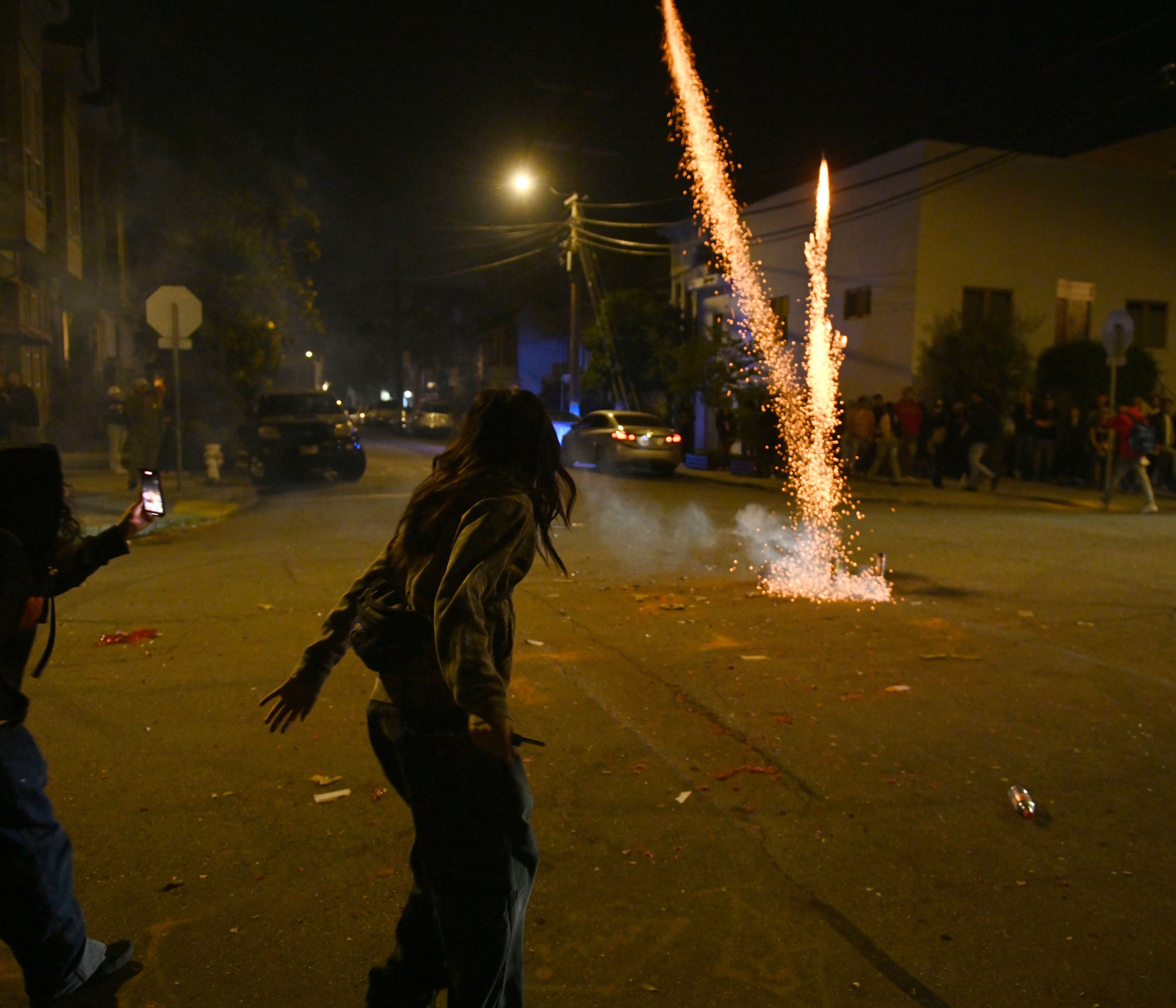 A street scene at night shows fireworks launching, with spectators watching, some recording on phones. The area is dimly lit with smoke, light, and scattered debris.