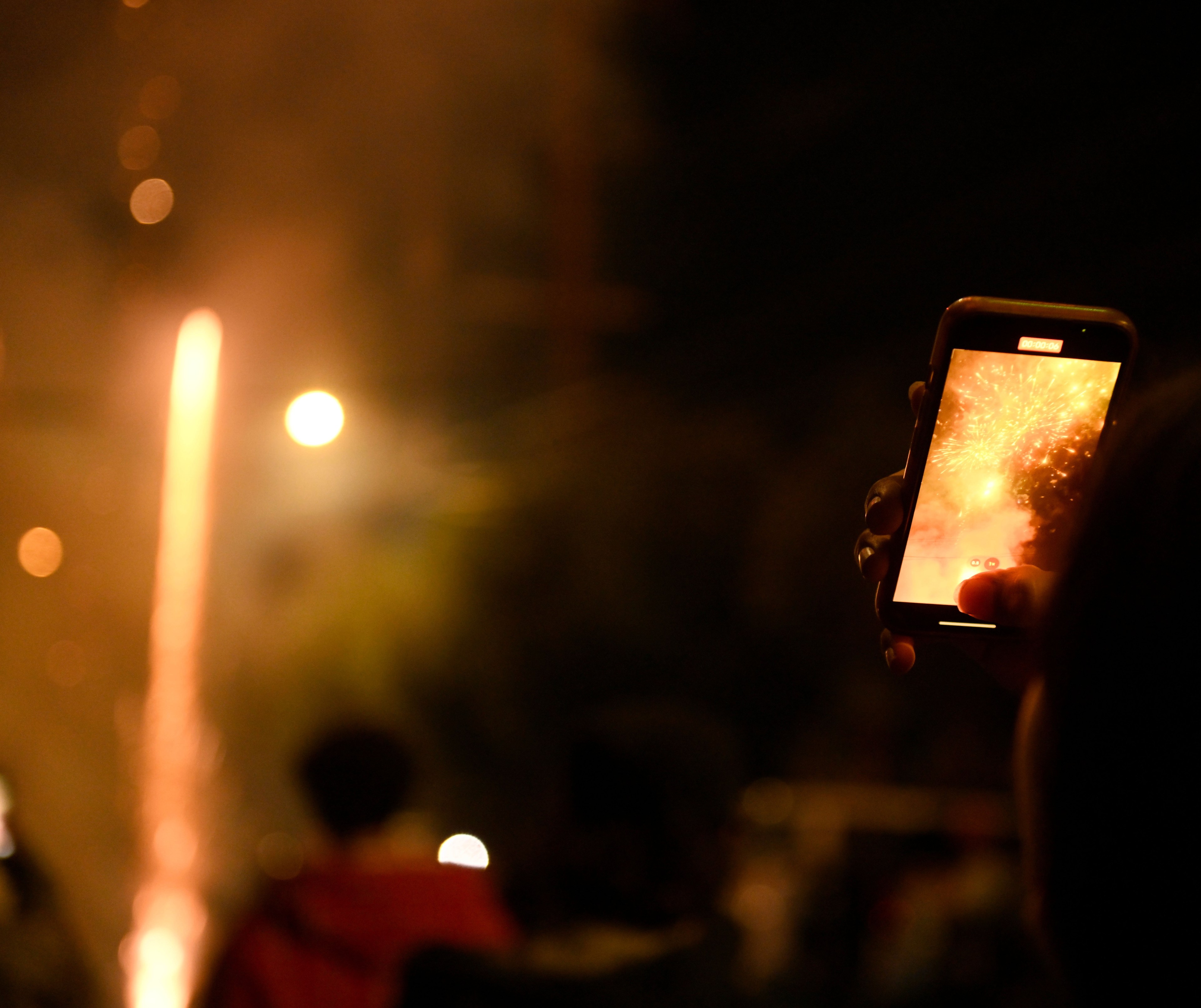 A person holds a phone, capturing fireworks in the night sky. The scene is brightly lit with fireworks, while the background is filled with slightly blurred lights and people.