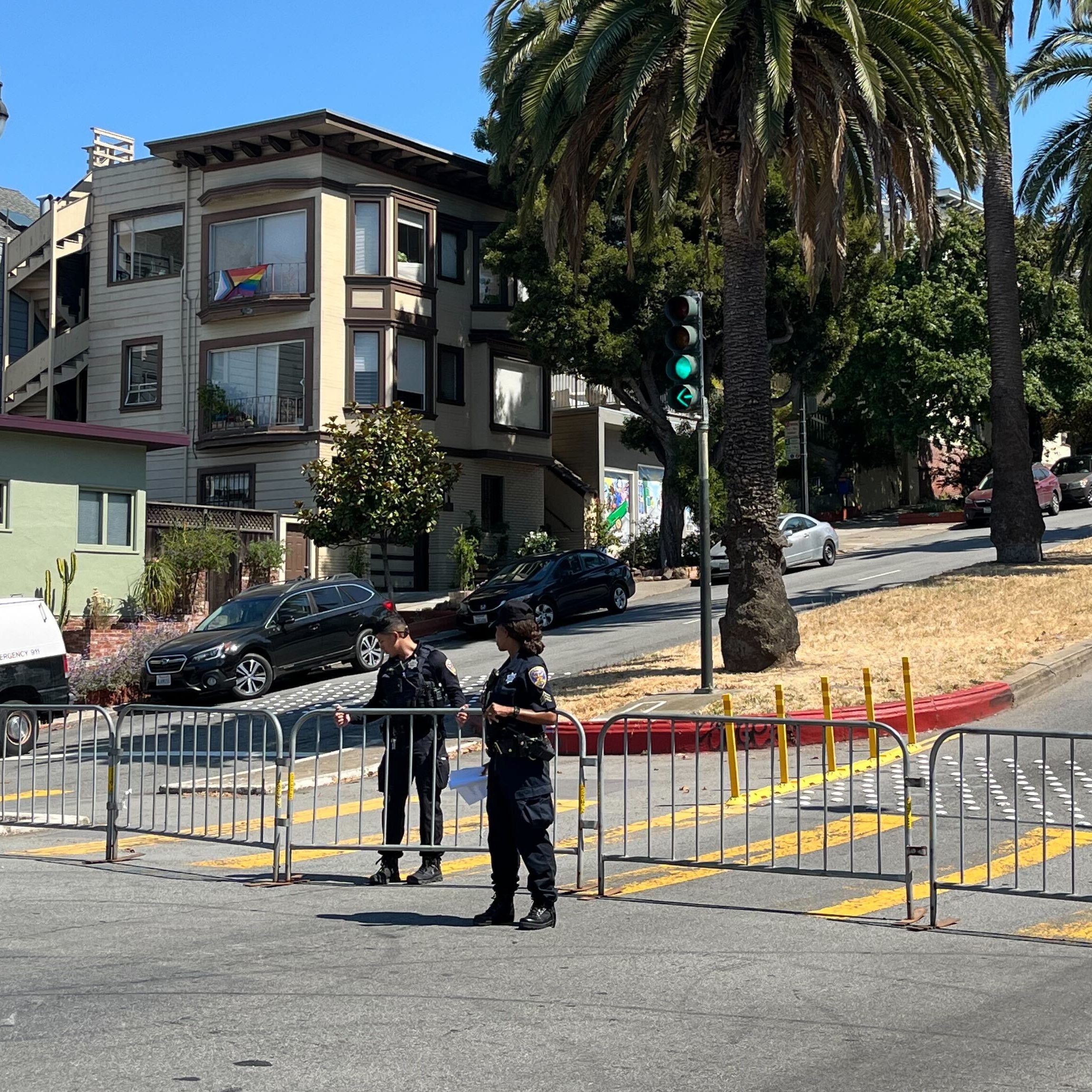 The image shows a street with police officers and barricades blocking traffic. Palm trees line the road, and houses are in the background under a clear blue sky.