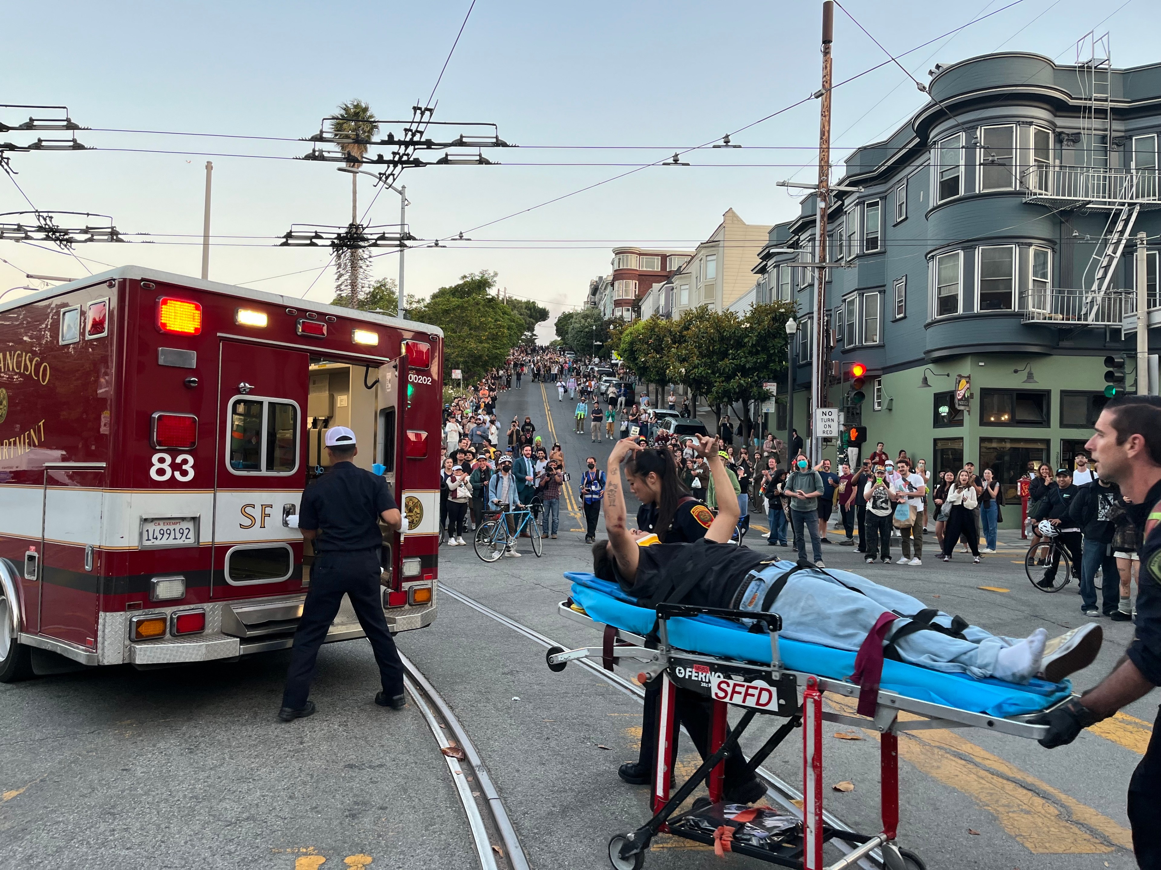 A person is being loaded into an ambulance on a stretcher by paramedics in a city street, while a crowd of onlookers watches from the sidewalk.