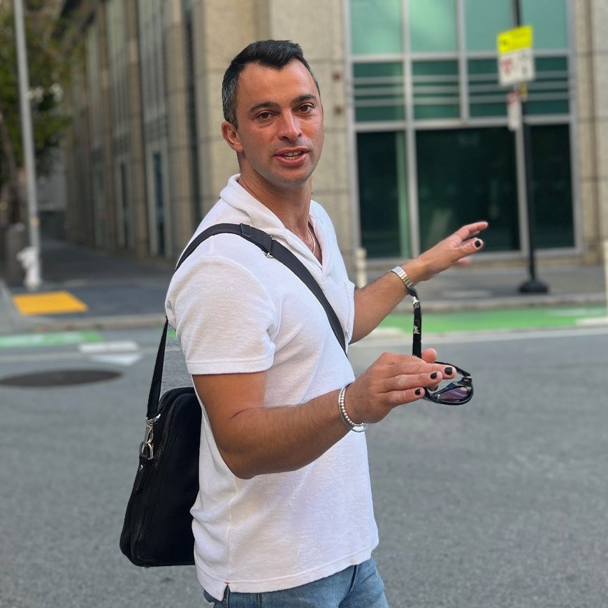 A man holding sunglasses stands in a city street, with a blurred woman in the foreground.