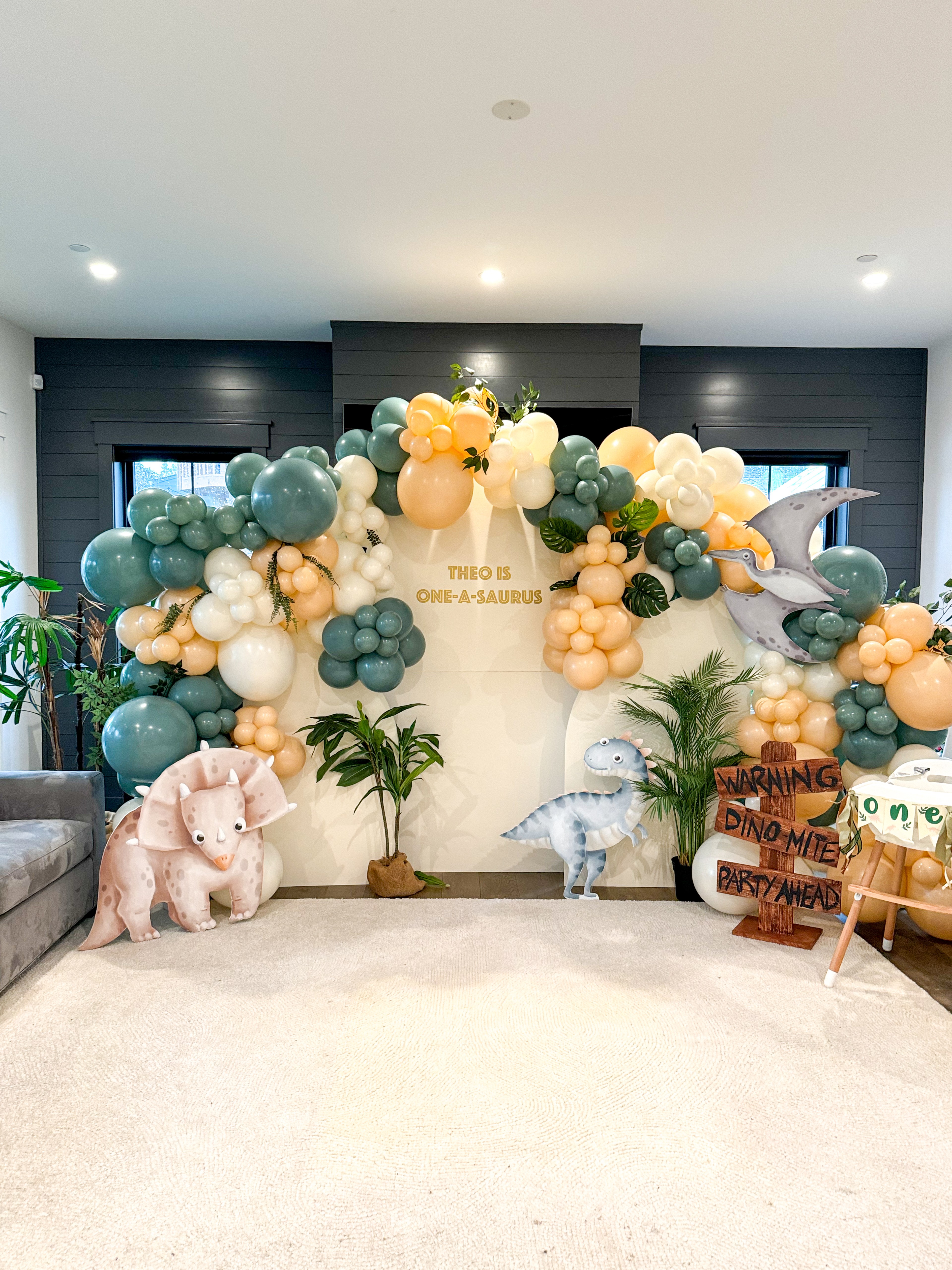 A dinosaur-themed party decor with a balloon arch, dinosaur cutouts, and a sign saying &quot;Theo is One-a-saurus.&quot; There are tropical plants and a wooden sign that reads &quot;Warning Dino-mite Party Ahead.&quot;