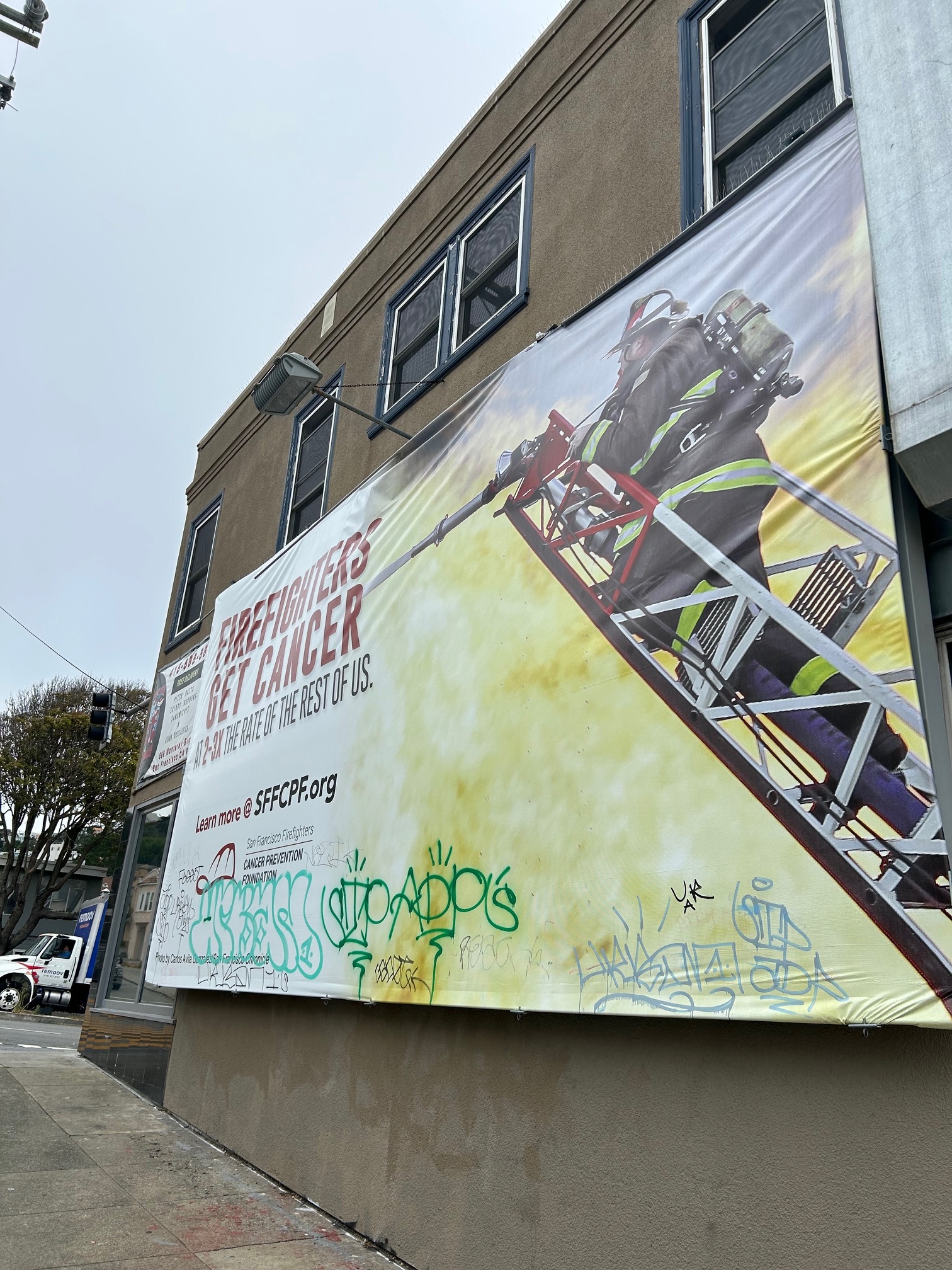 The image shows a large banner on a building's exterior, advocating for firefighter cancer prevention; part of it is covered in colorful graffiti. A firefighter is pictured on a ladder.