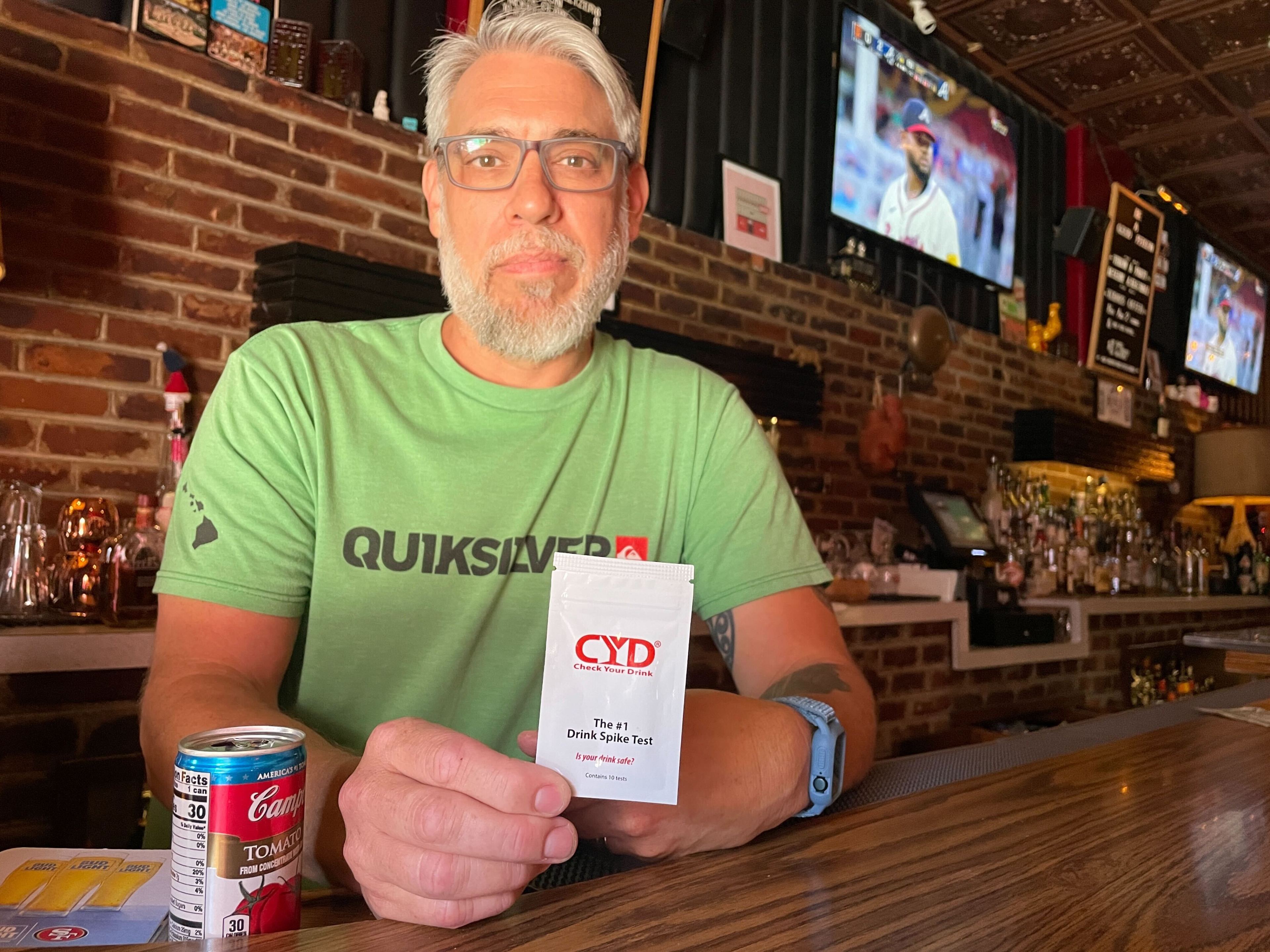 A man stands behind a bar holding a white package that says "Drink Spike Test" on the front.