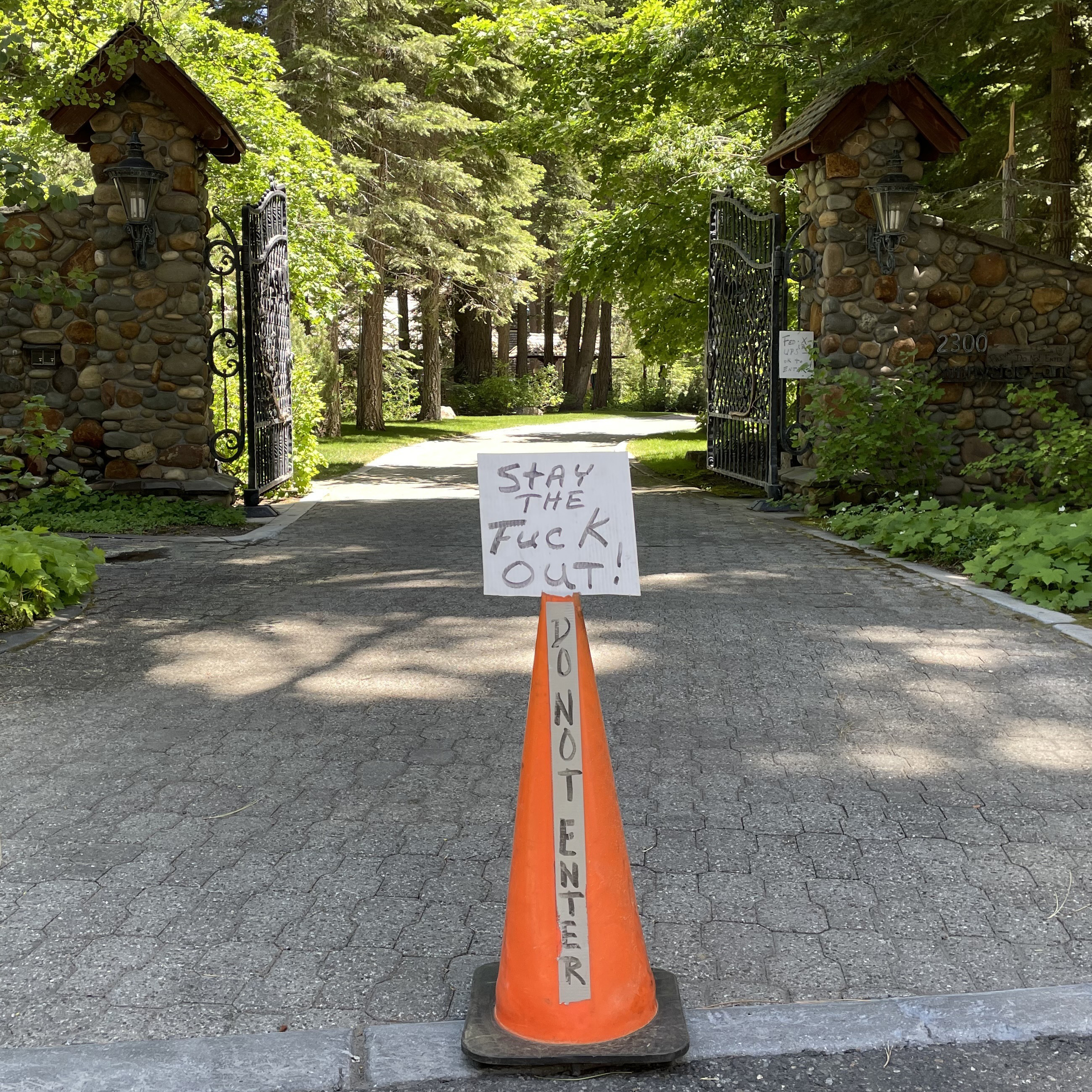 The image shows a gated driveway with a stone entrance, and a traffic cone bearing a sign that reads, "STAY THE FUCK OUT!" with "DO NOT ENTER" written vertically on the cone.